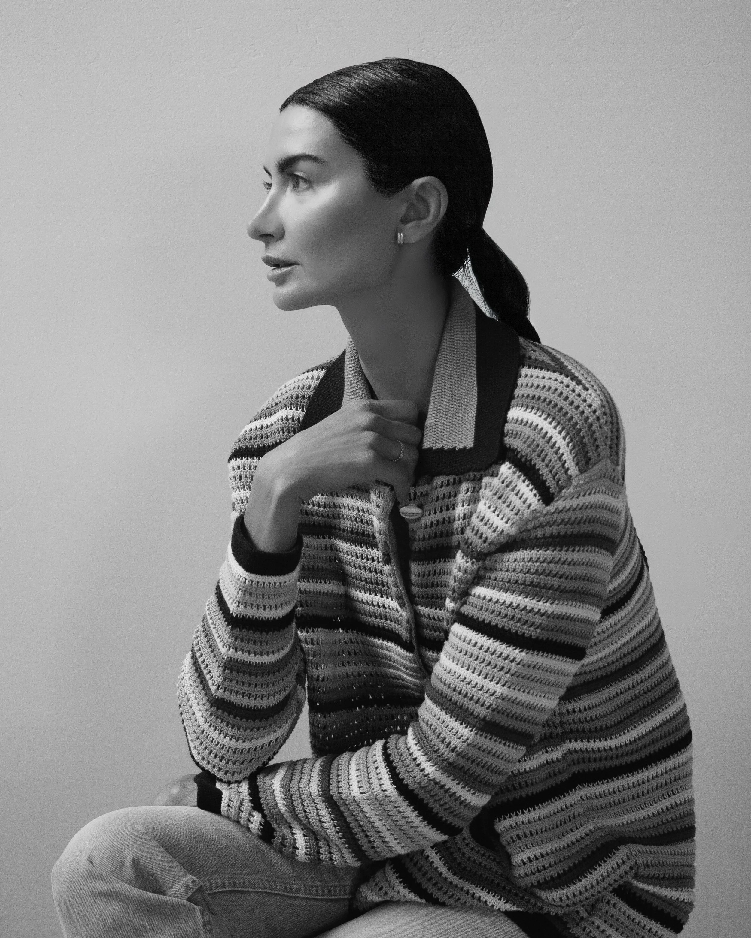 A black and white photo of a woman with dark hair pulled back, wearing a striped sweater, sitting with one arm crossed over her knee and her hand near her neck, against a plain wall background.
