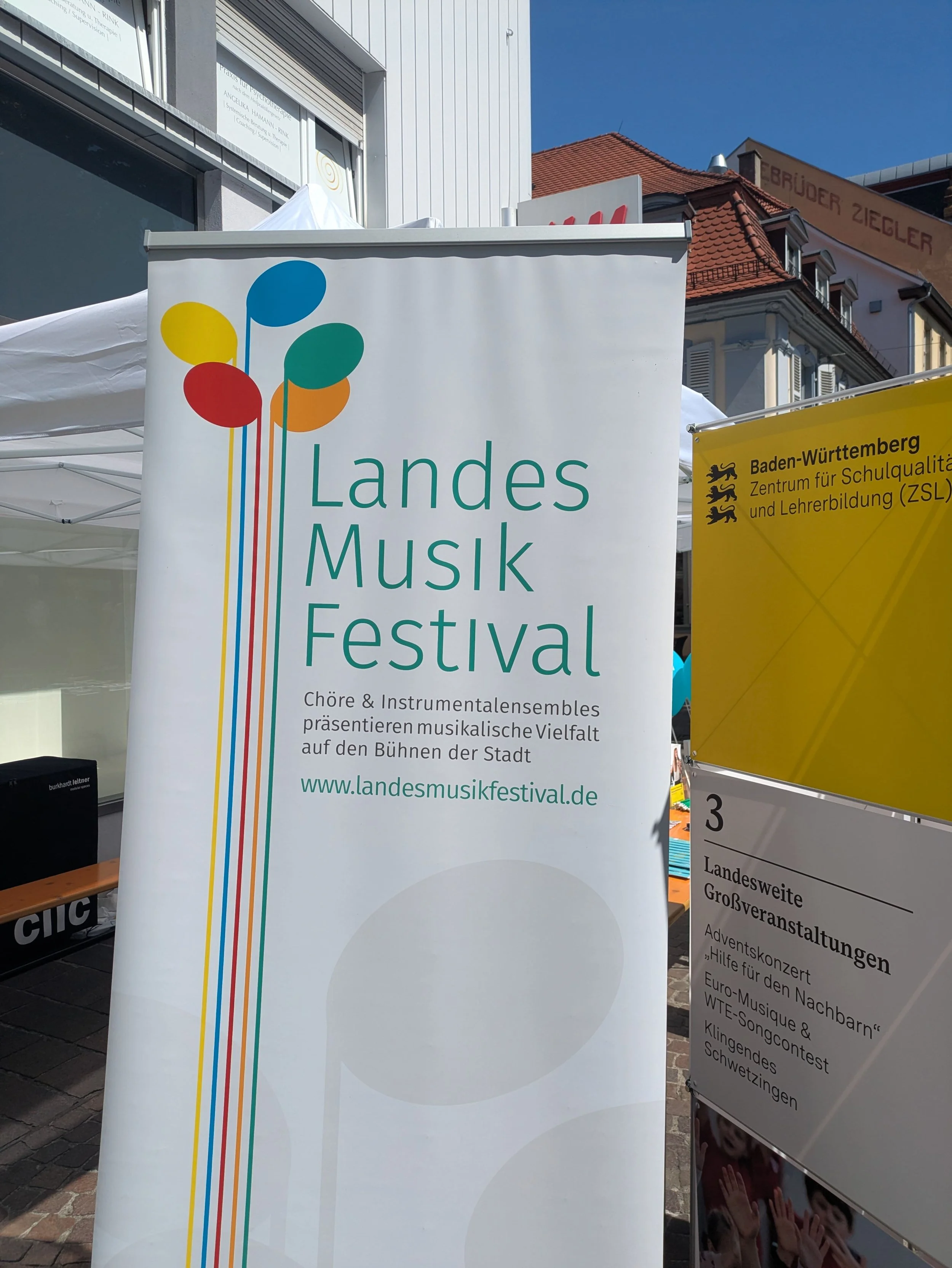 Ein weißer Plakataufsteller mit der Aufschrift Landesmusikfestival und dem Logo der Veranstaltung. Das Logo besteht aus bunten Noten, die wie ein Blumenstrauß angeordnet sind.