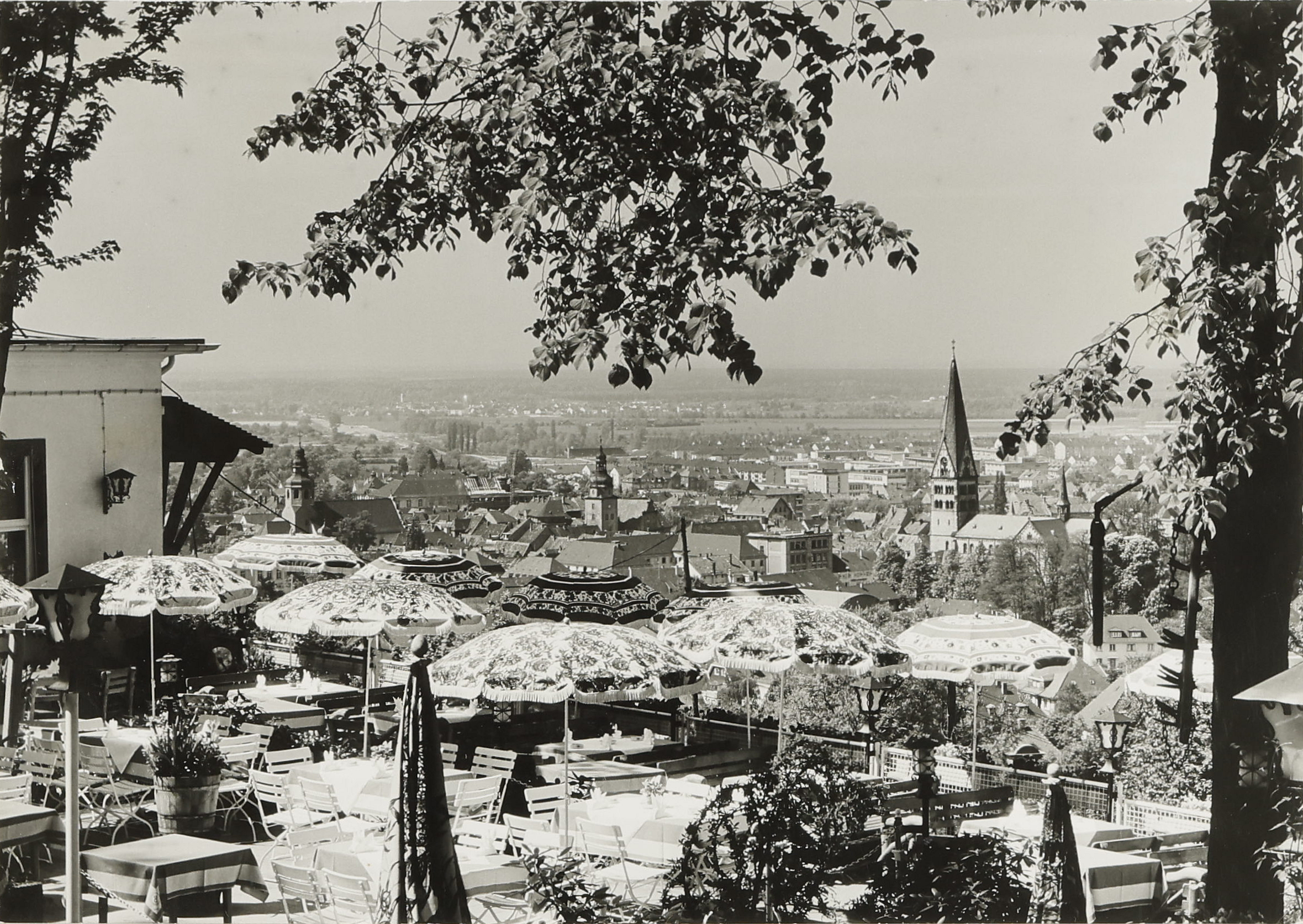 Ein schwarz-weiß Bild, auf dessen Hintergrund eine Stadt zu sehen ist. Im Vordergrund ist eine Terrasse mit Sonnenschirmen.