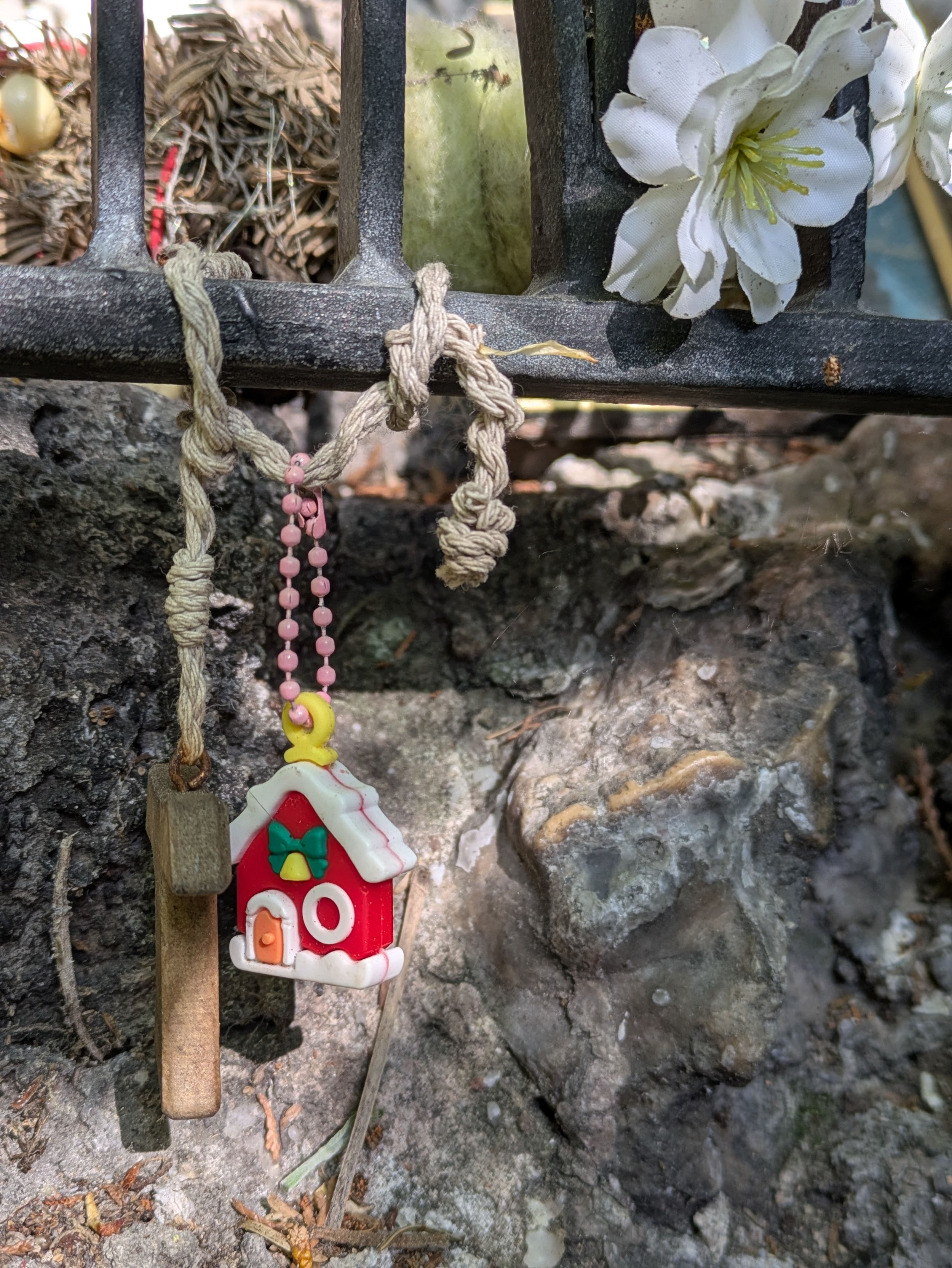 Zwei Anhänger, die am Gitter vor der Mutter Gottes hängen: ein Holzkreuz und ein kleines rotes Haus mit weißem Dach.