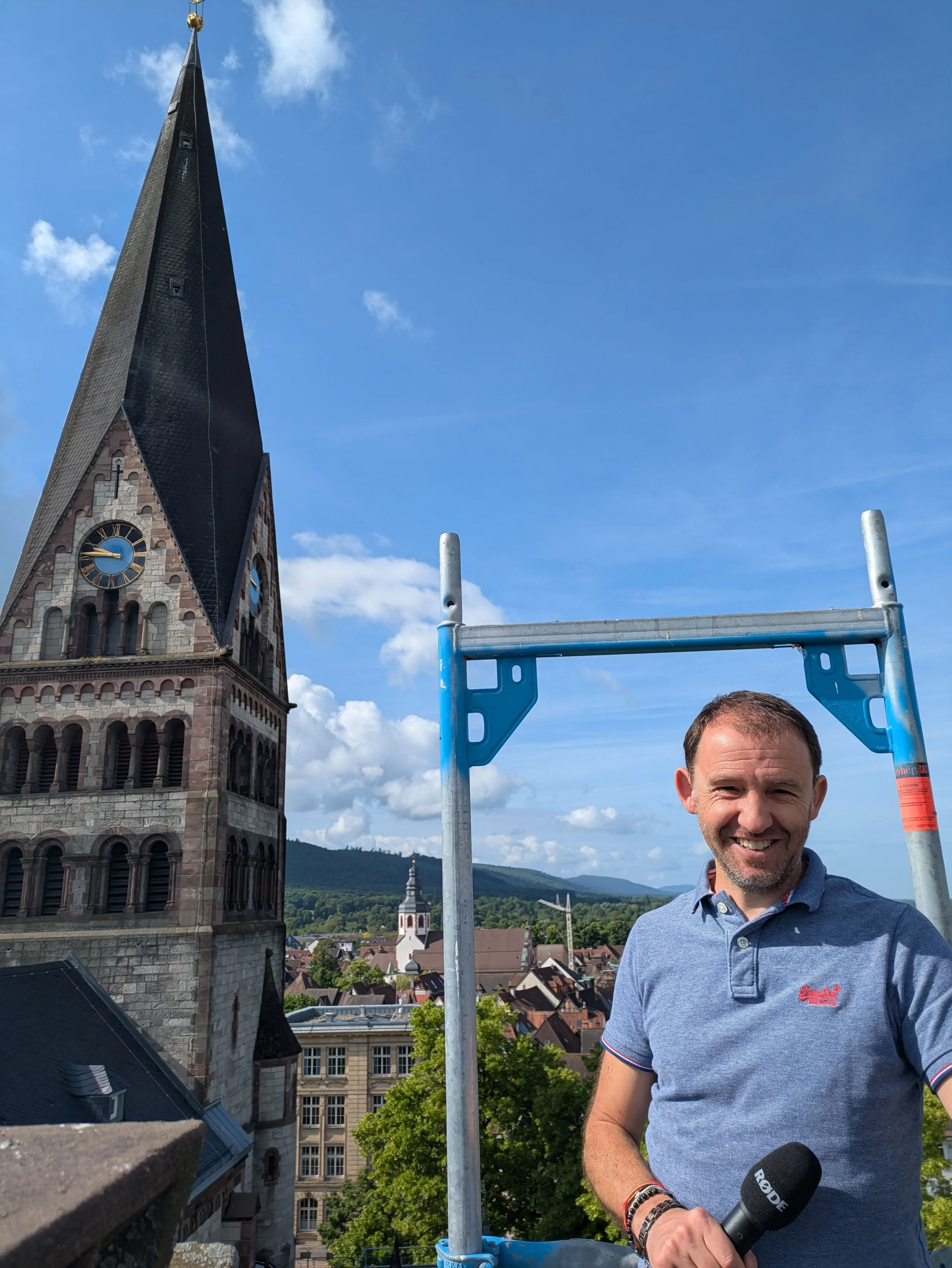 Ein Mann mittleren Alters in blauem T-Shirt und mit Mikrofon in der Hand auf einem Gerüst an einer Kirche.