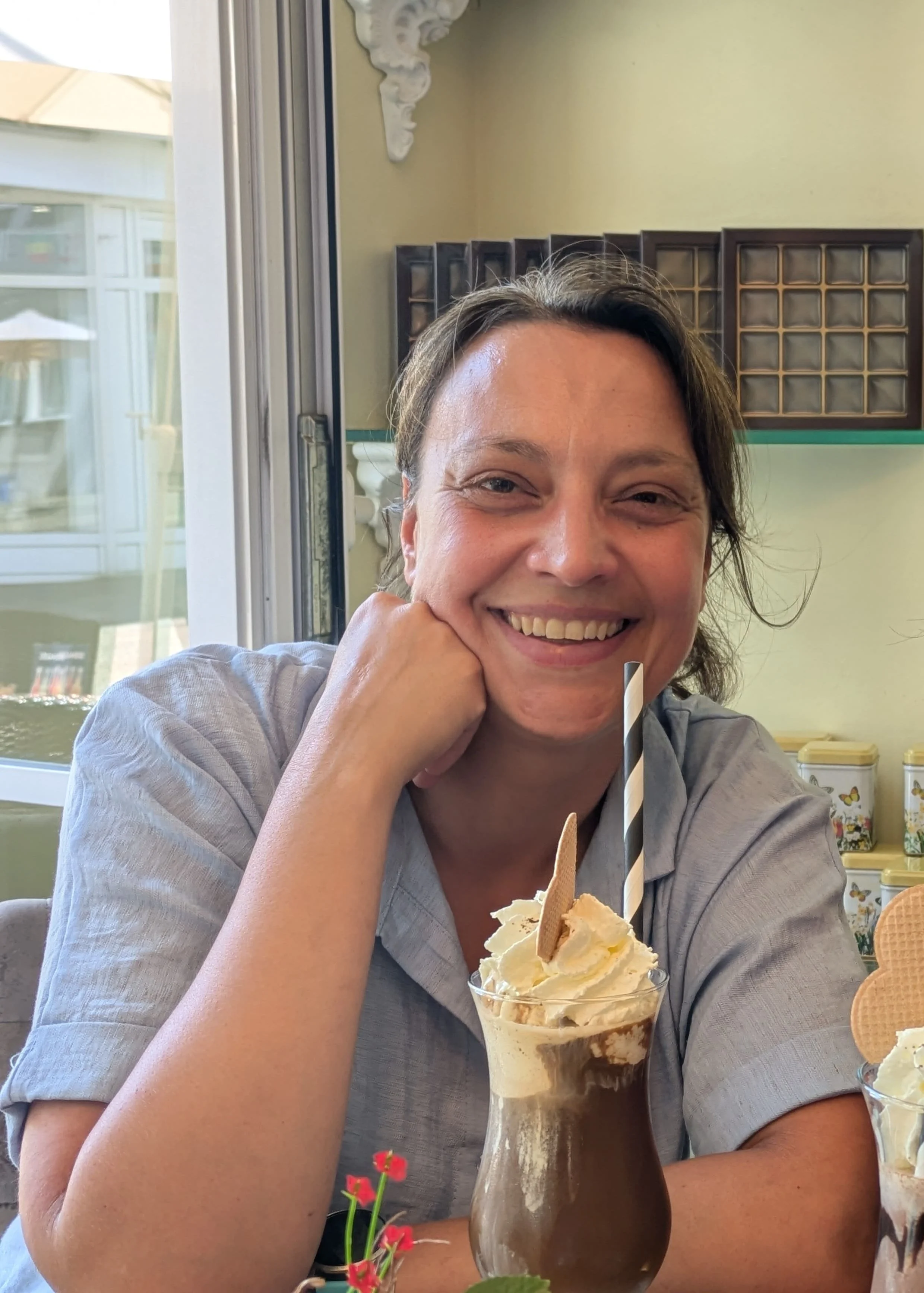 Eine Frau mittleren Alters mit braunen Haaren, die zu einem Pferdeschwanz gebunden sind. Sie lächelt in die Kamera. Vor ihr steht ein Eiskaffee mit viel Sahne.