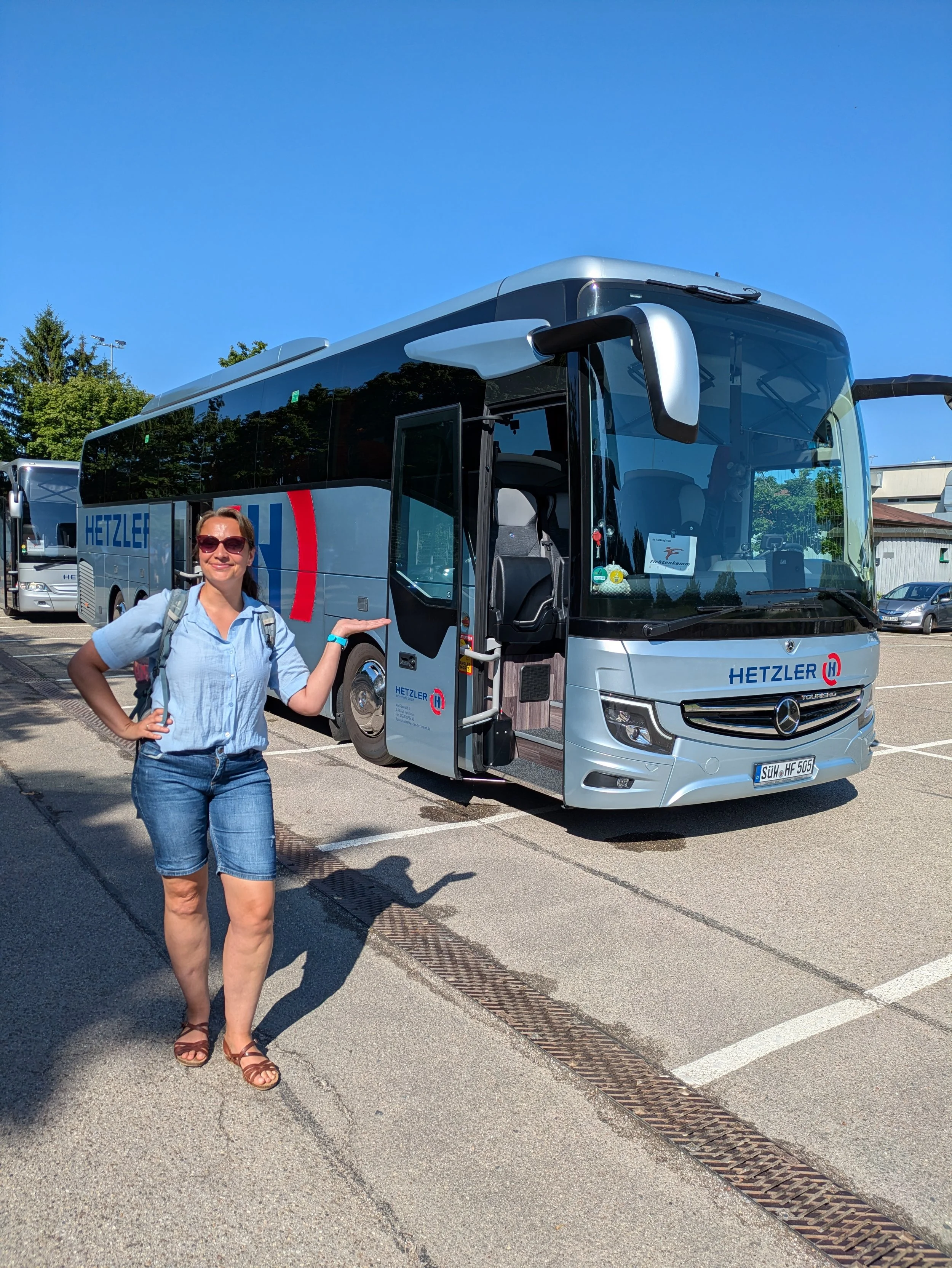 Eine Frau mittleren Alters, gekleidet in Jeans und hellblauer Bluse, steht vor einem Reisebus.