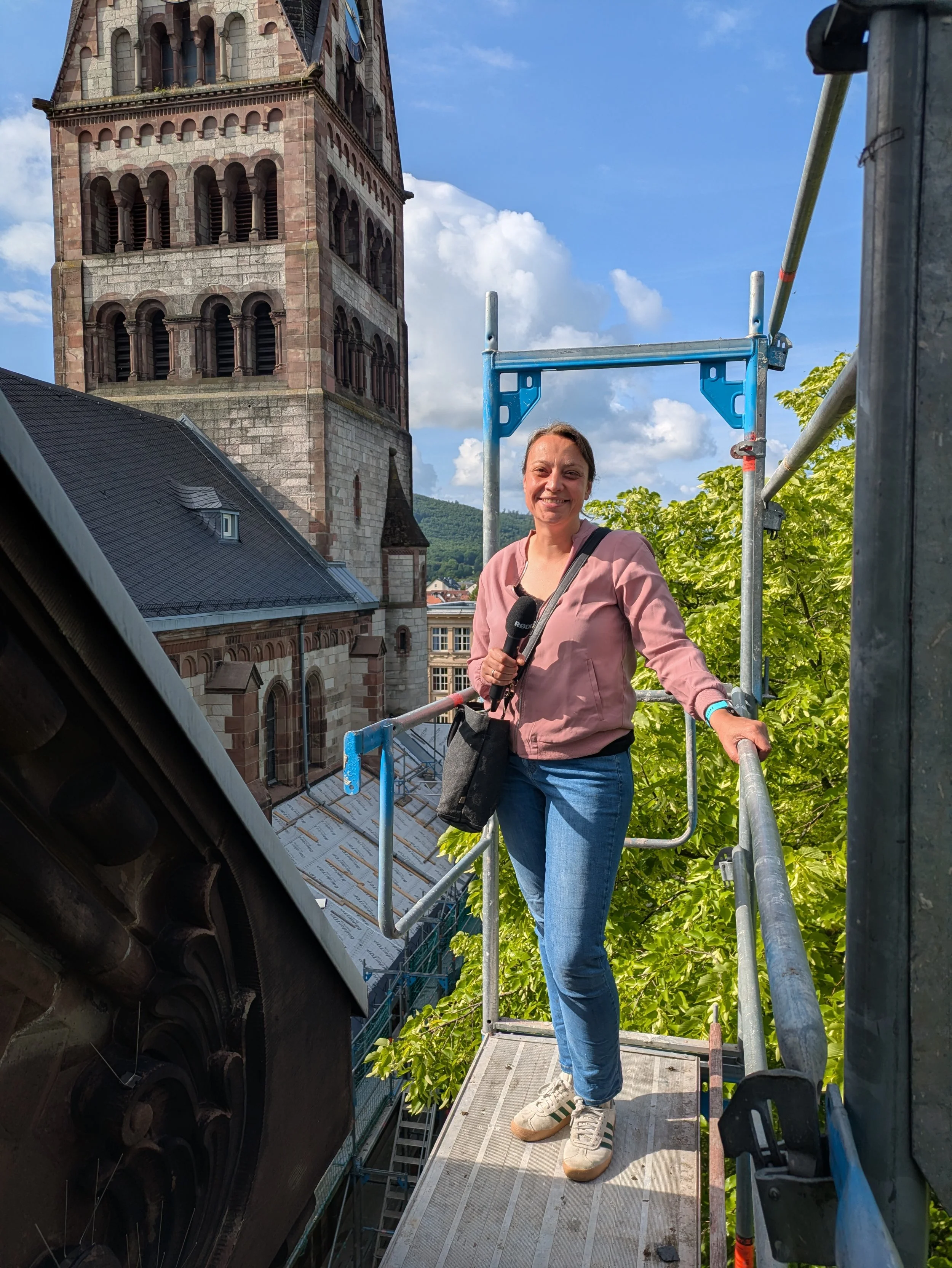 Eine Frau mittleren Alters in Jeans, rosa Pullover und Mikrofon in der Hand auf einem Gerüst an einer Kirche.