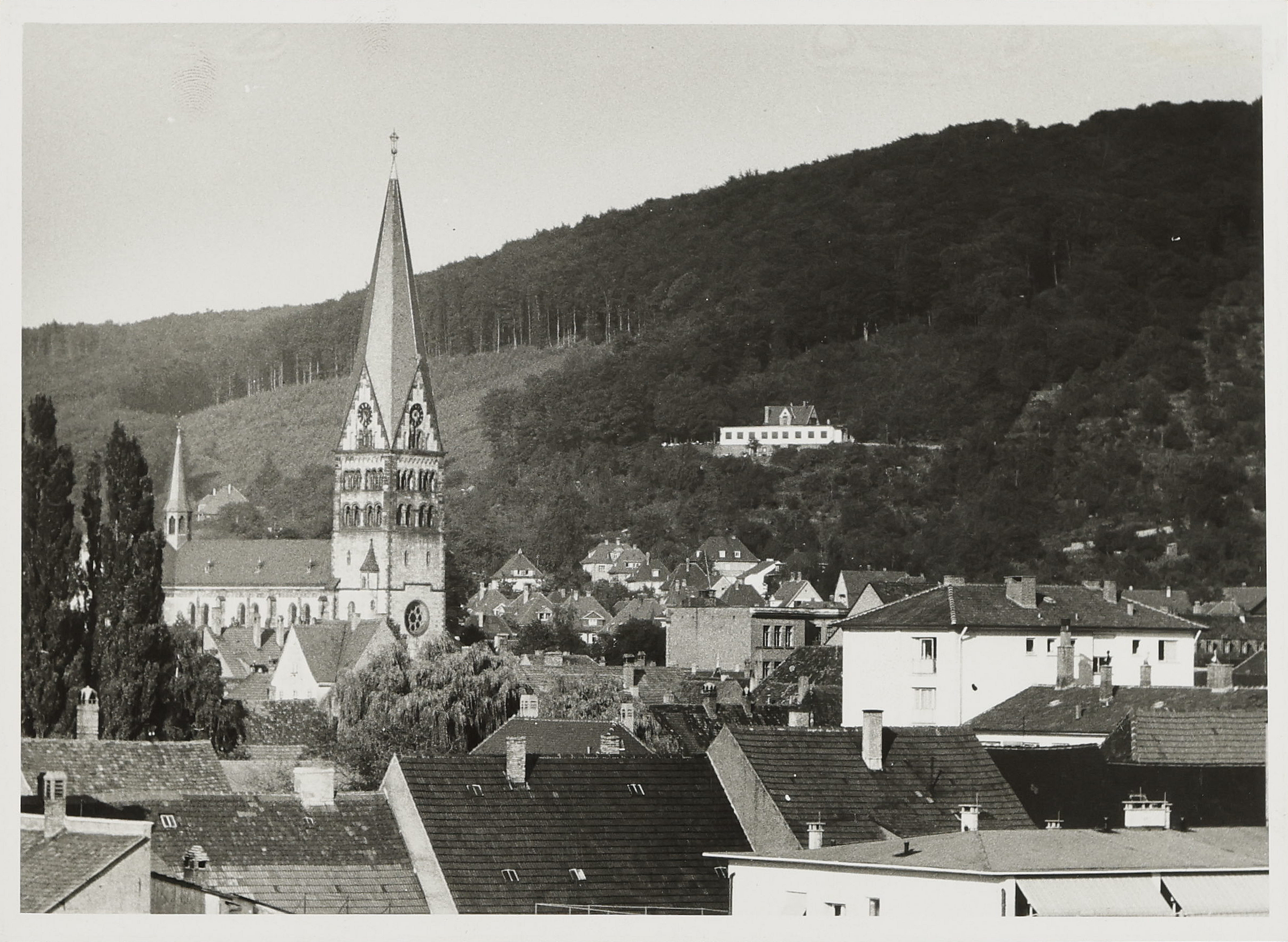 Ein schwarz-weiß Bild mit einer Stadt im Vordergrund, aus der auf der linken Seite ein Kirchturm empor ragt. Im Hintergrund sieht man einen bewaldeten Hand, an dem ein kleines, weißes, flaches Gebäude zu sehen ist.