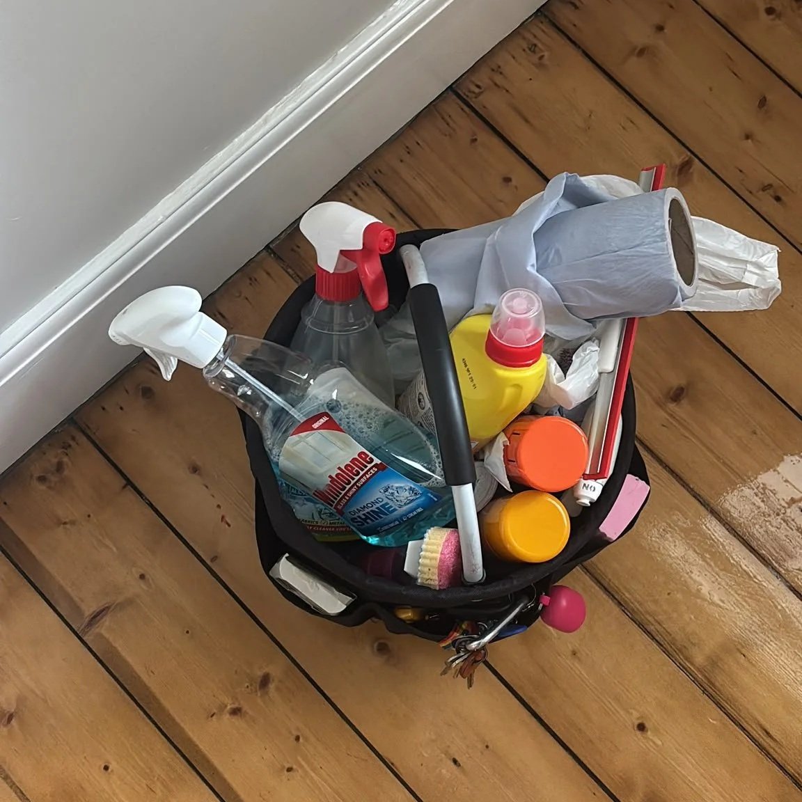 A small black laundry basket filled with cleaning supplies, including spray bottles, a sponge, a scrub brush, and plastic containers, placed on a wooden floor near a white wall and baseboard.