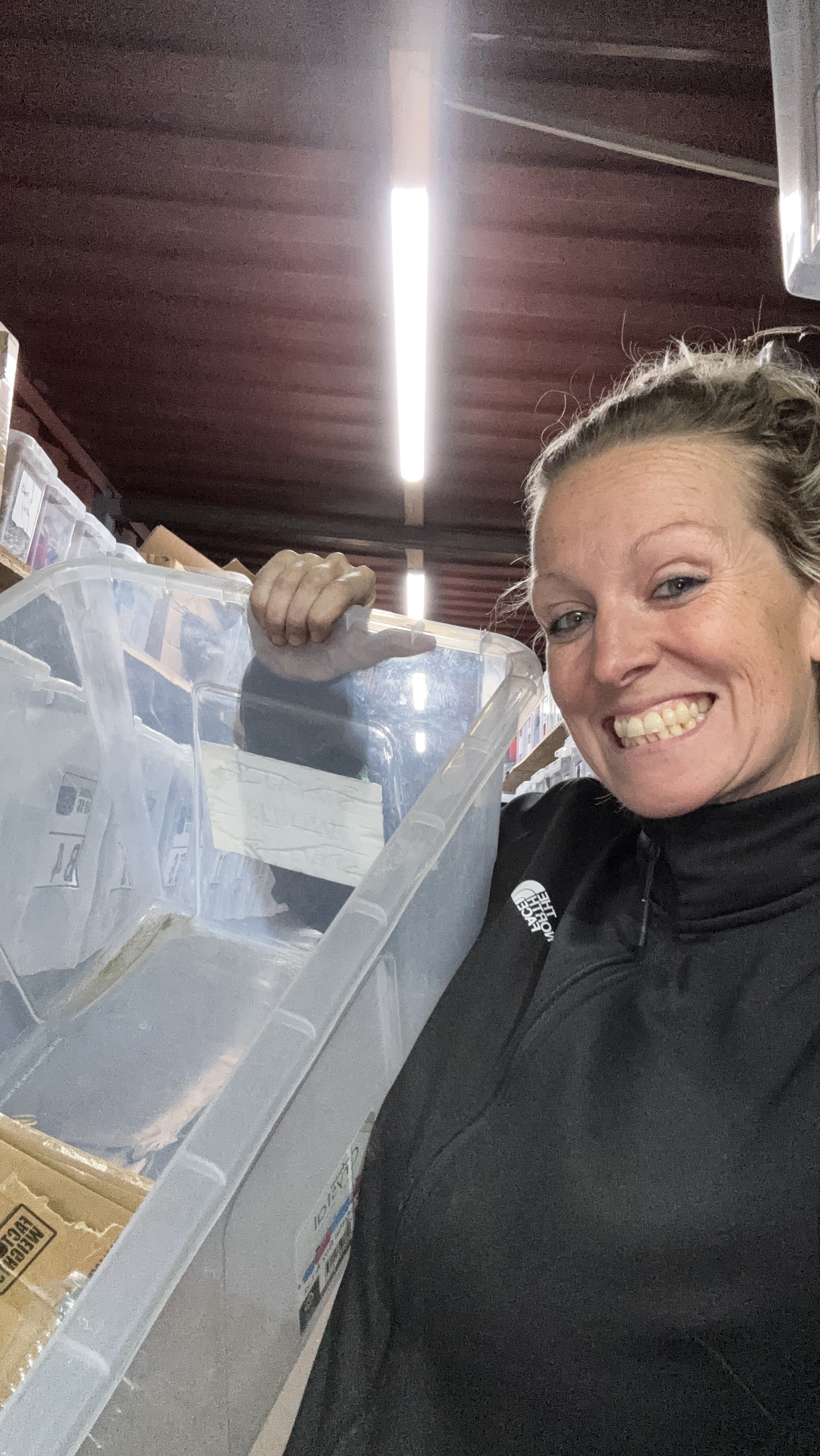 A woman smiling and holding a plastic storage container filled with various items in a storage or warehouse space with shelves and fluorescent lighting.