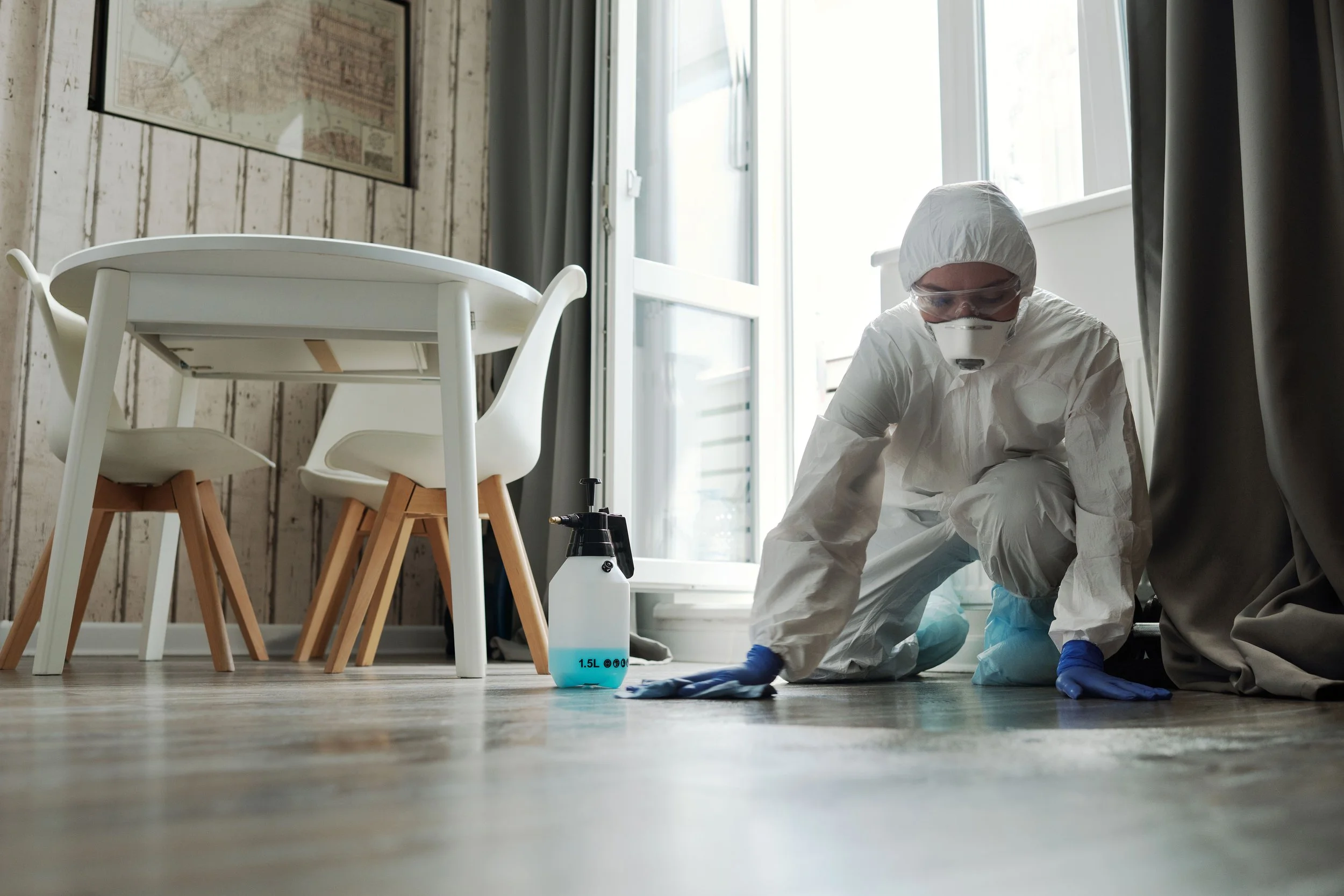Person in protective gear cleaning the floor with a cloth and spray bottle in a bright room.