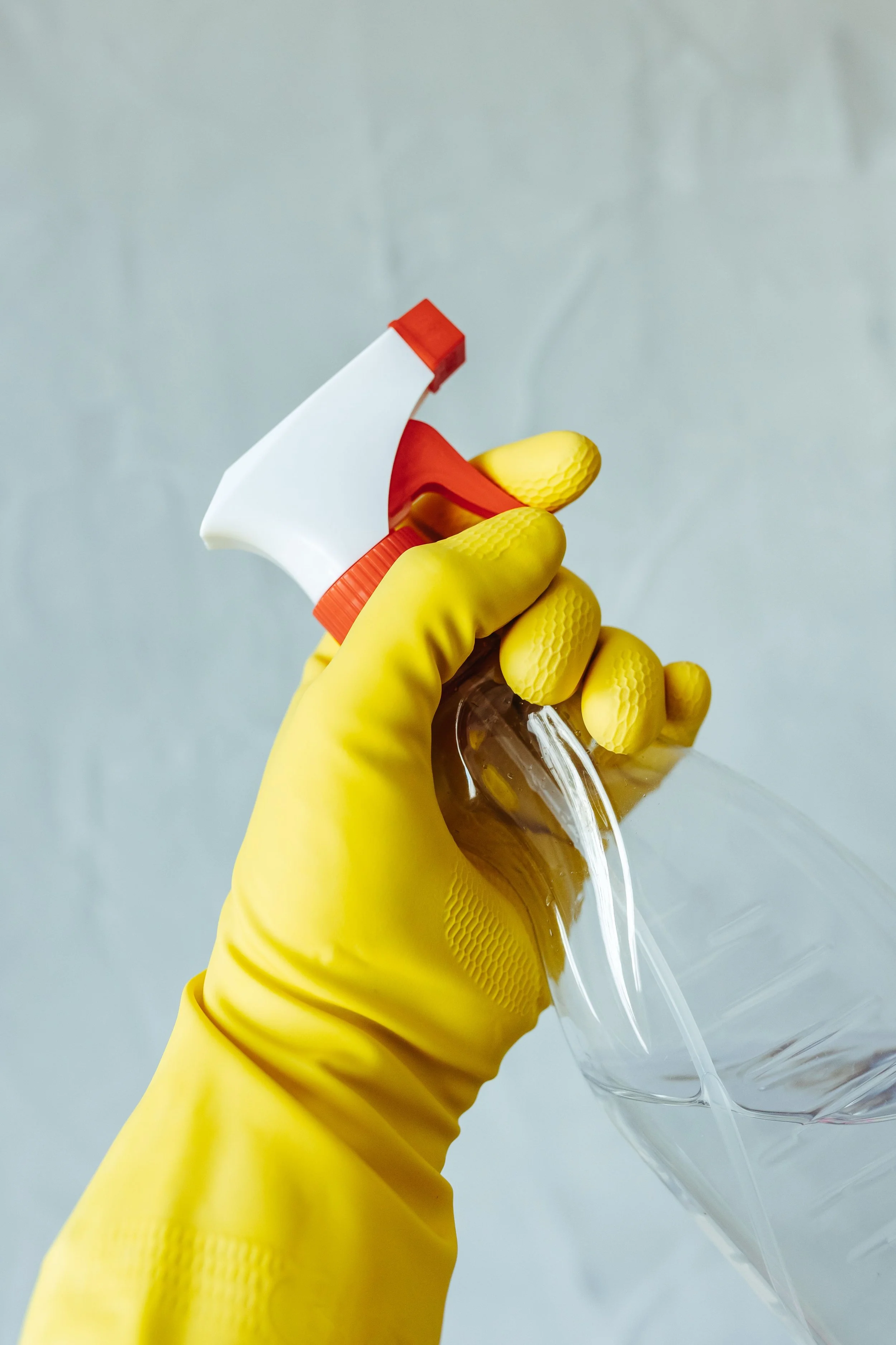 Person wearing yellow gloves holding a spray bottle and pouring a liquid into a clear container against a light gray background.