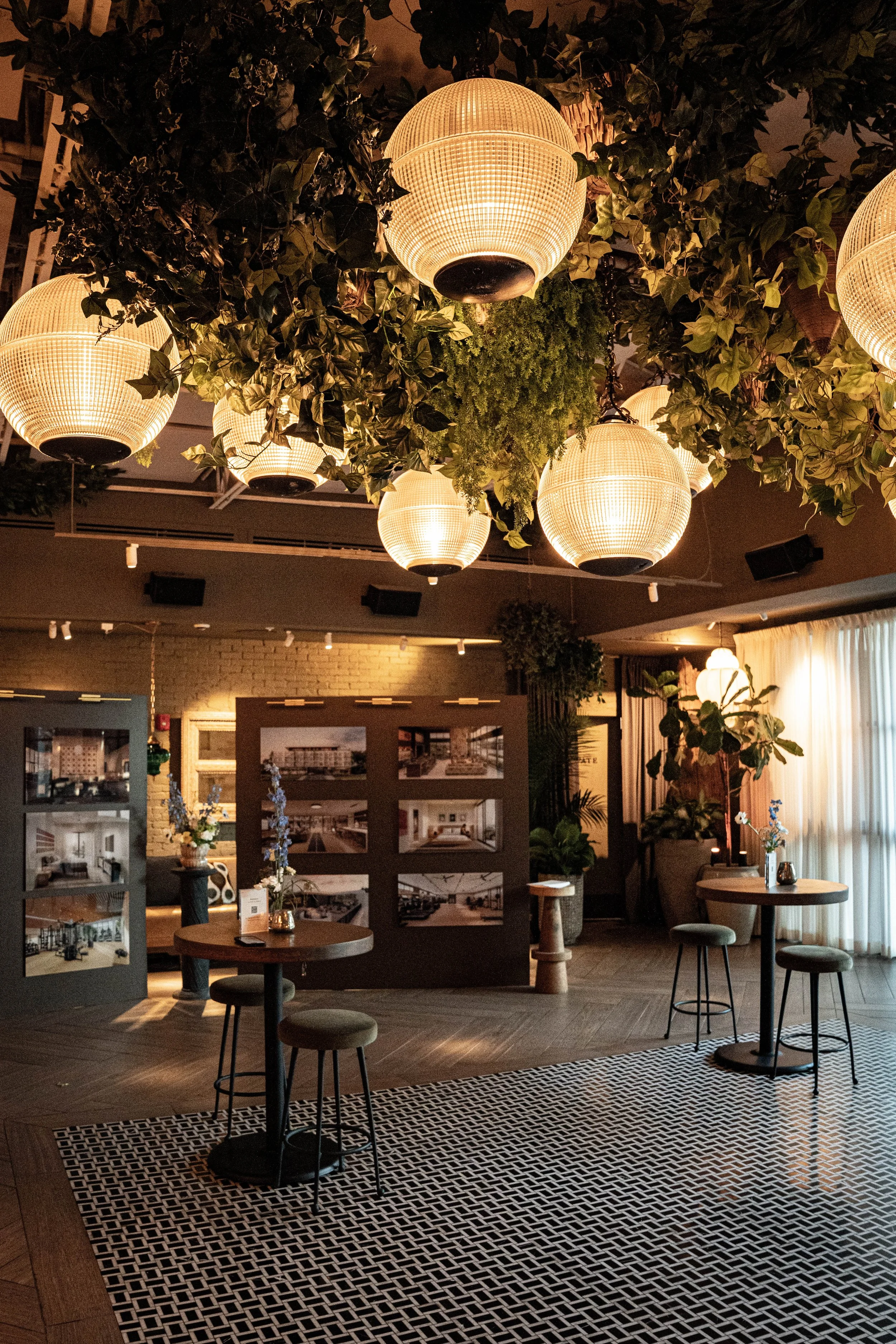 Interior of a modern room with hanging paper lanterns, green plants, and photo displays on a partition. Small tables with stools and chairs are arranged around. Warm, ambient lighting creates a cozy atmosphere.