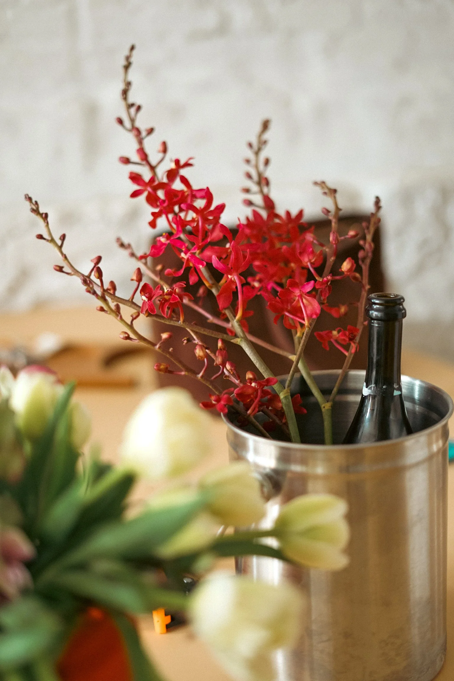 A red flowering plant in a brown pot placed inside a metallic container with a black bottle near it, with a blurred background of a beige wall.