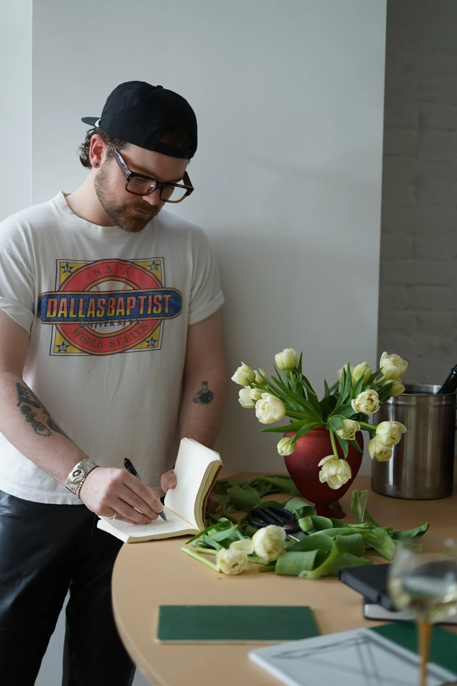 A man with glasses and a backwards cap is writing in a notebook at a table, surrounded by white tulips in a red vase and cutting shears.