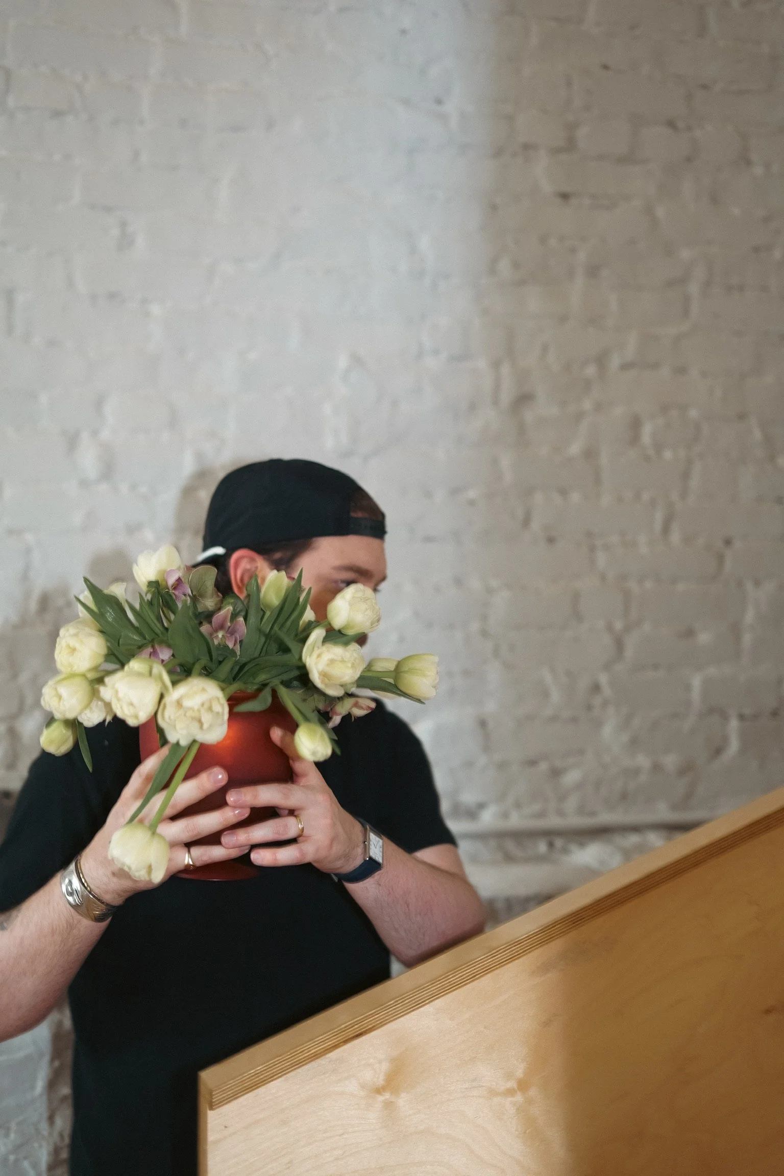 A person with a baseball cap holding a pot of white and pink tulips, partially covering their face, in front of a brick wall.
