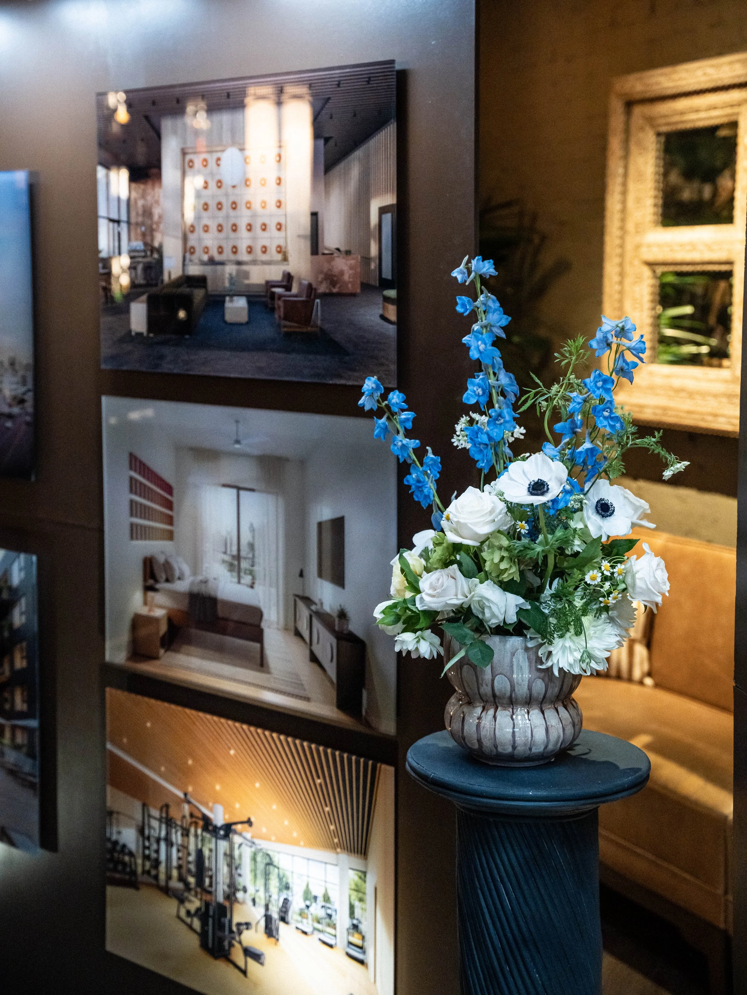 A photograph of a gallery wall showcasing interior design images with a bouquet of blue and white flowers in a decorative vase on a black ornate pedestal in the foreground.