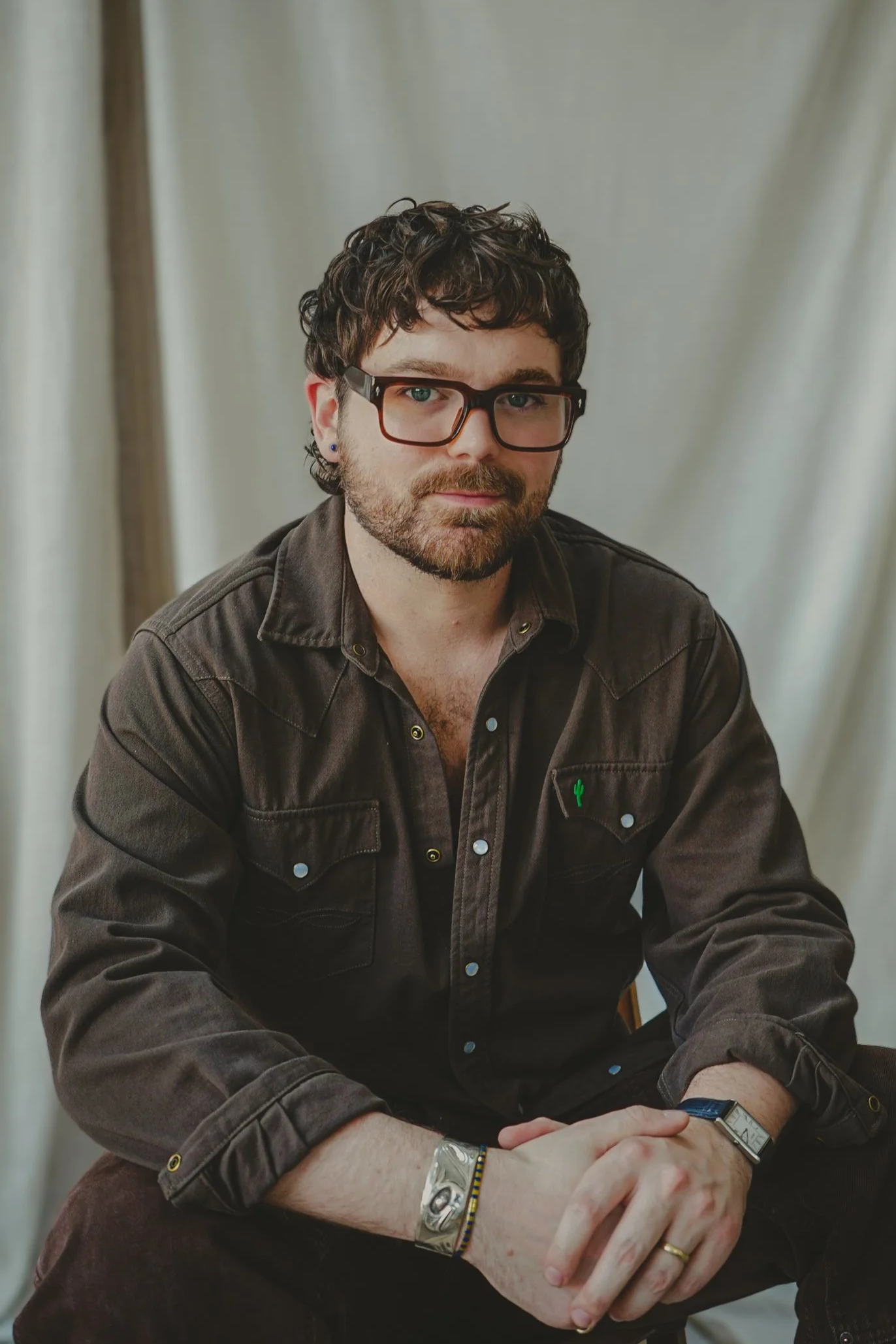 A young man with curly brown hair, glasses, and a beard, wearing a dark brown denim shirt, sitting in front of beige curtains, with his hands crossed on his lap. He has multiple bracelets, a wristwatch, and a ring.