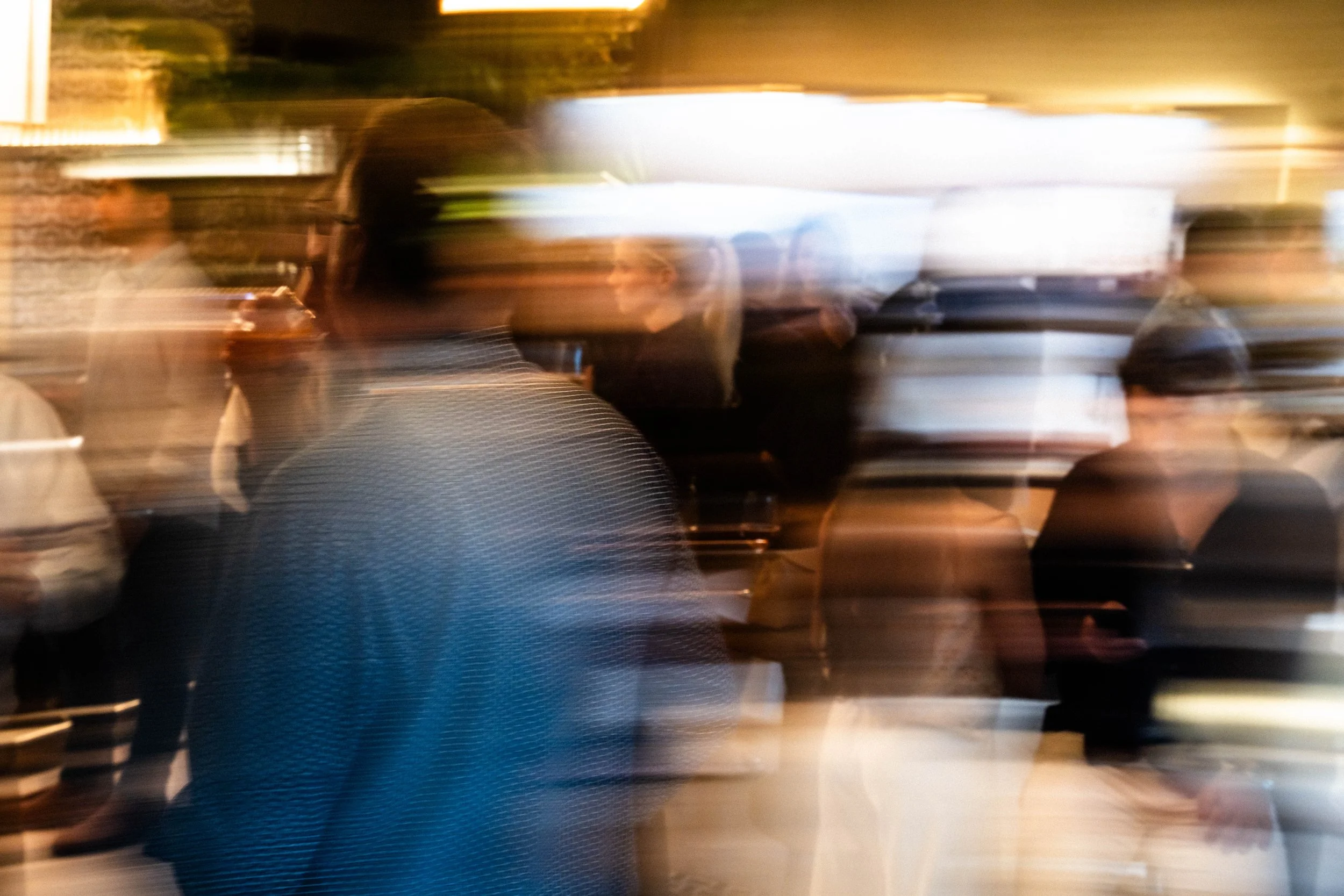 Blurry photo of several people in a crowded indoor setting, possibly a restaurant or event space, with bright lighting and motion blur.