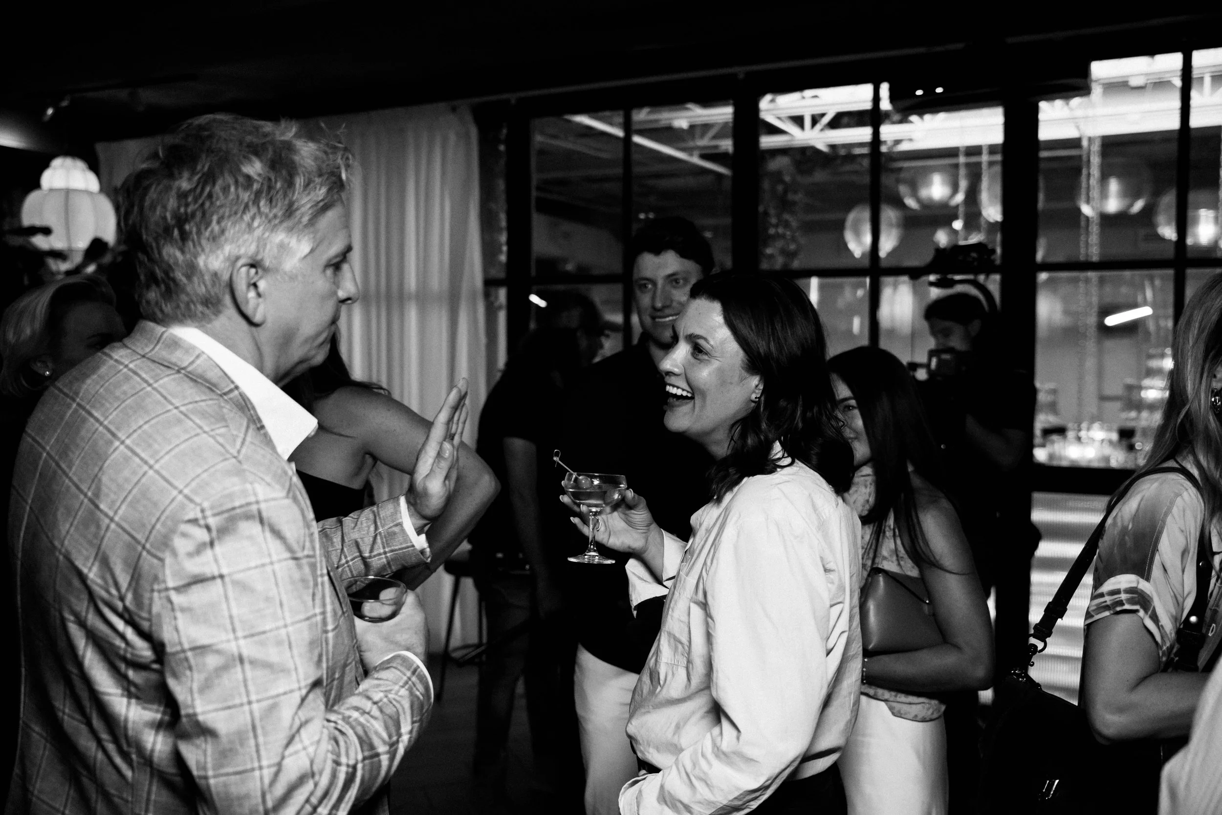 People socializing and chatting at a party or gathering indoors, with large windows and decorative lighting in the background.