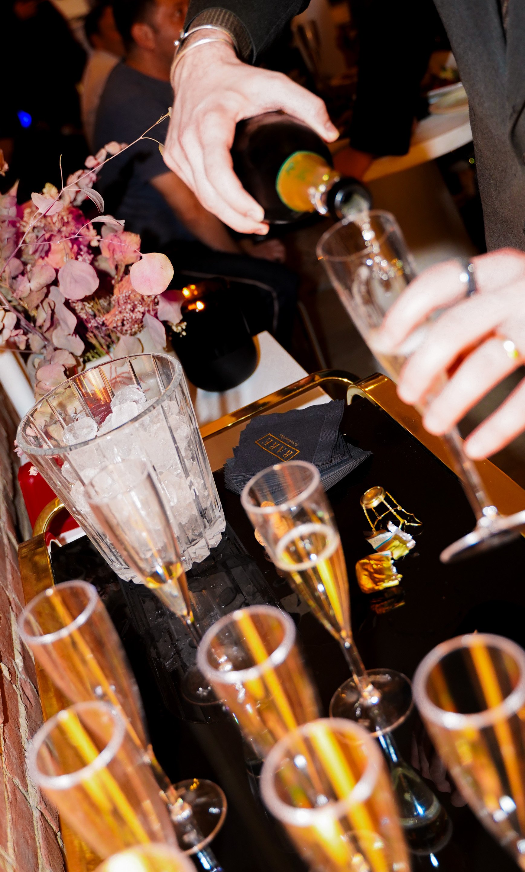 A person pours a dark bottle of champagne into a tall flute glass at a celebration event, with multiple champagne glasses on a black tray, a bouquet of pink and purple flowers, and people in the background.