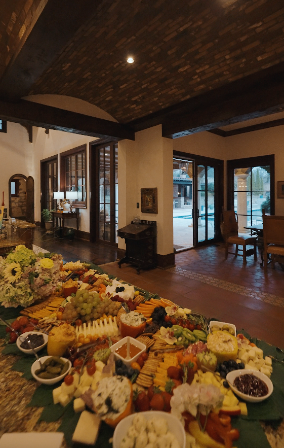 Cheese and fruit platter with various cheeses, grapes, crackers, and garnishes on a dining table in a rustic living room.