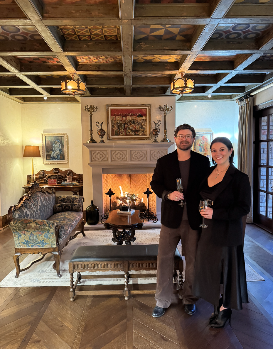 A man and woman stand together in a cozy, vintage-style living room with a smiling expression, holding champagne glasses. The room features a decorative fireplace with artwork above, antique furniture, and warm lighting, creating an elegant atmosphere.