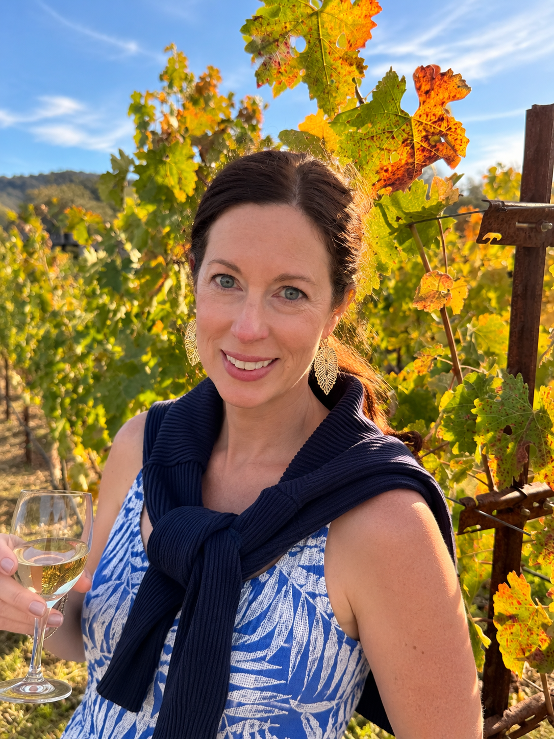 Shay Palms founder styled in a palm print dress with an Alo sweatshirt tied over the shoulders on a crisp autumn day among vineyard fall foliage