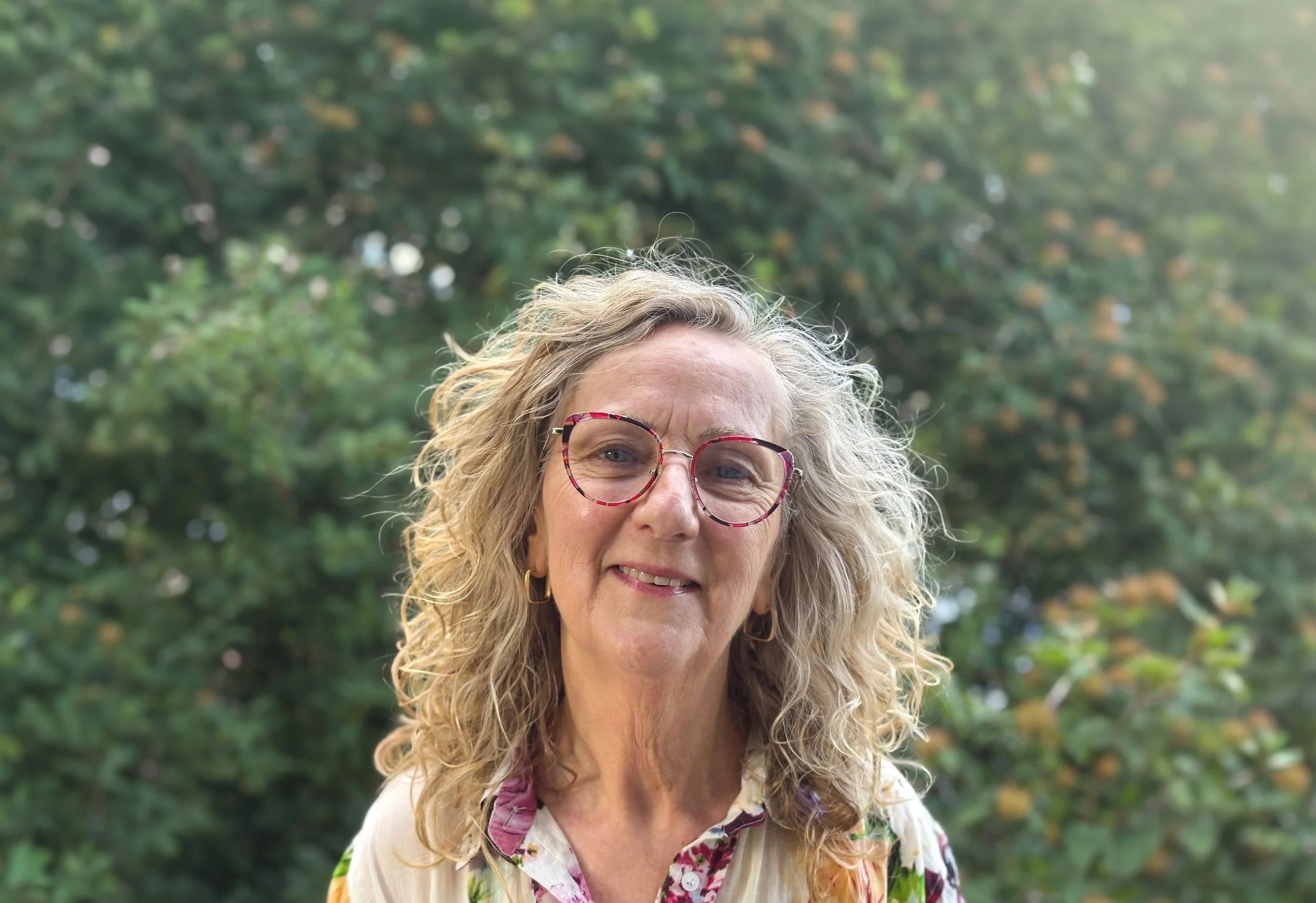 Close-up of a woman with curly hair, wearing patterned glasses, smiling outdoors with a blurred green leafy background.