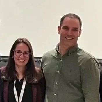 A woman and a man smiling together, standing indoors against a plain wall.