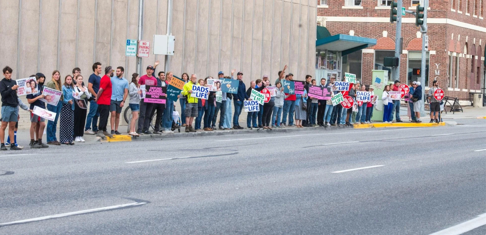 A large group of protesters stands in a line on a city sidewalk holding signs supporting pro-life causes and opposing abortion.