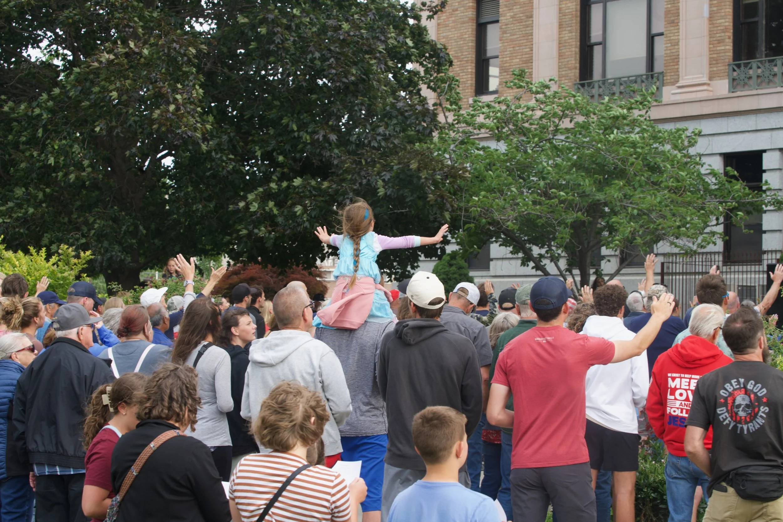 A large crowd of people gathered outdoors, with a young girl in a blue jacket and pink pants sitting on someone's shoulders among the crowd. Some people have their hands raised, and there are trees and buildings in the background.