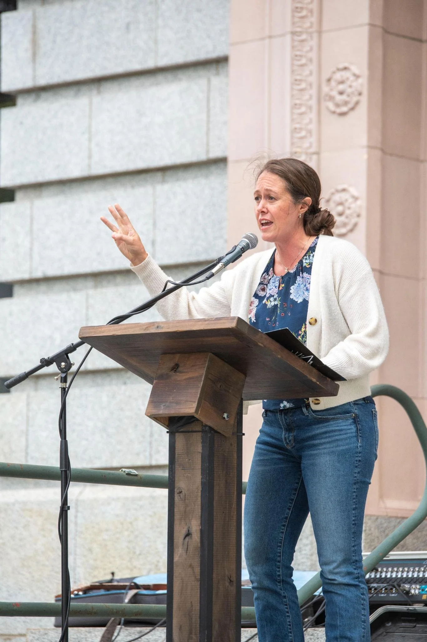 A woman with brown hair in a bun speaking at a podium outdoors, gesturing with her left hand while standing in front of a beige building with decorative architectural details.