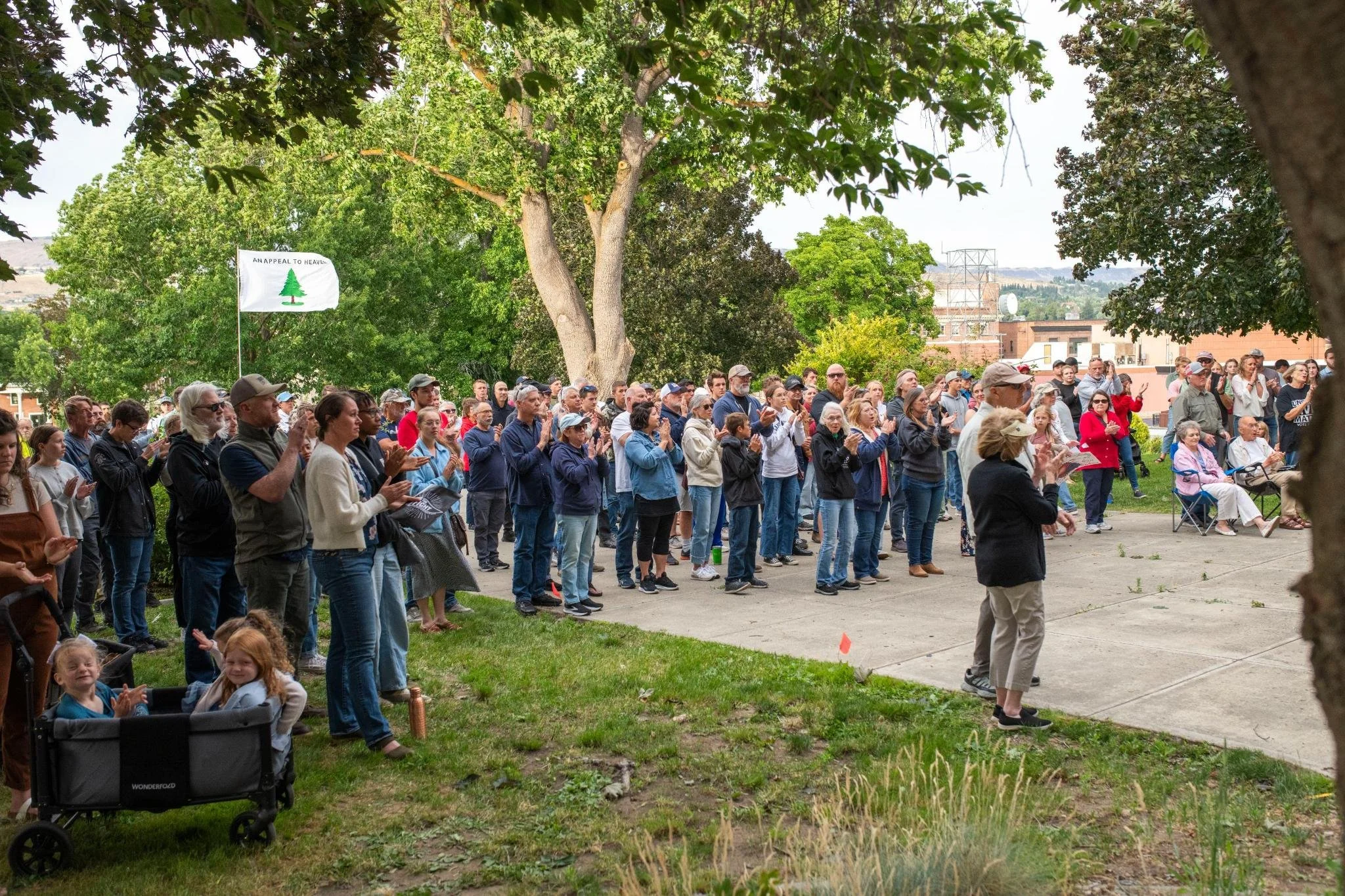 A crowd of people gathered outdoors, clapping and listening attentively, with trees and a building in the background.