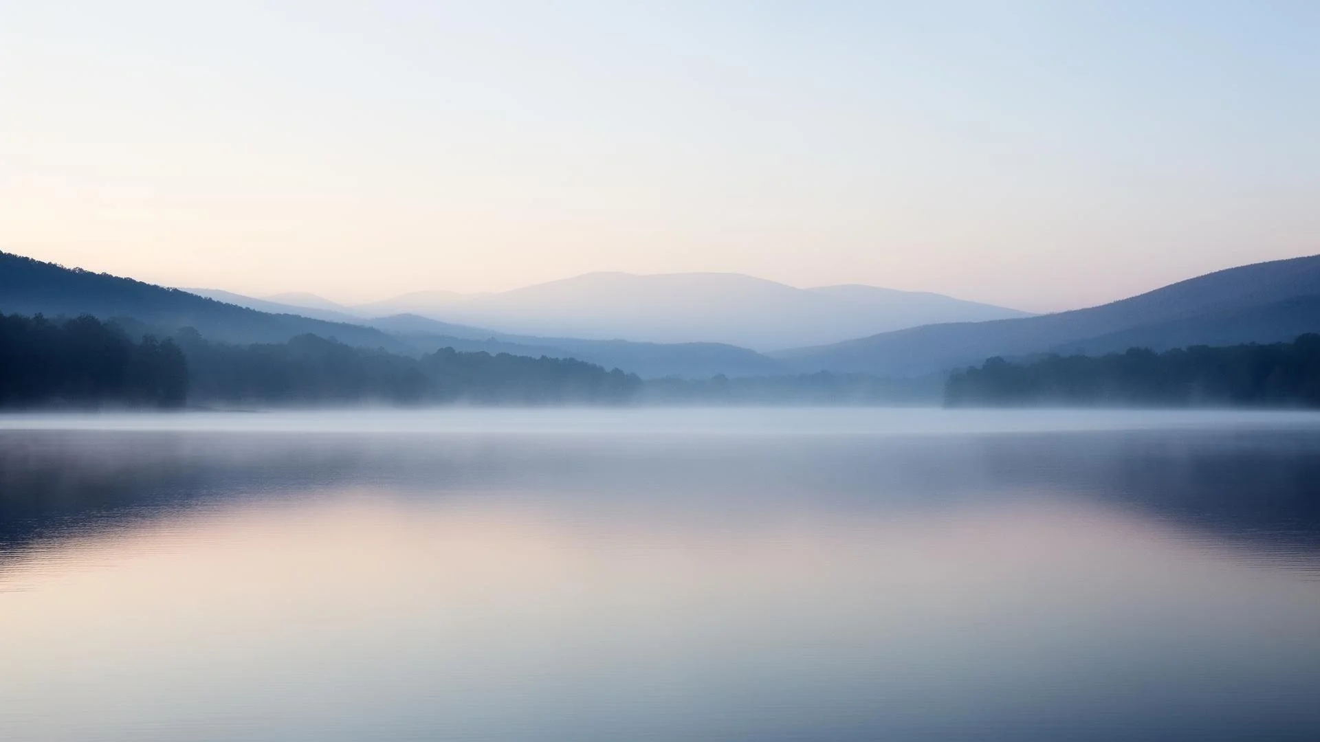 Calm lake with mist rising over the water, surrounded by mountains and forest in the distance, under a pastel-colored sky.