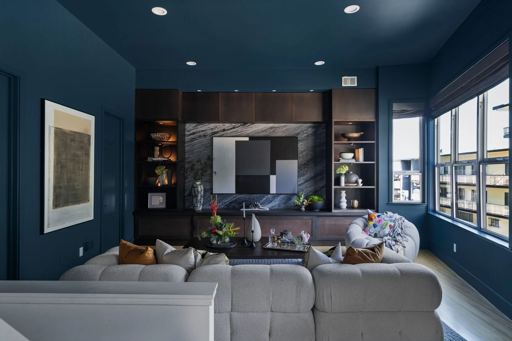 Living room with blue walls and ceiling, a gray couch with pillows, a black TV console with decorative objects, and a window with blinds revealing a building outside.
