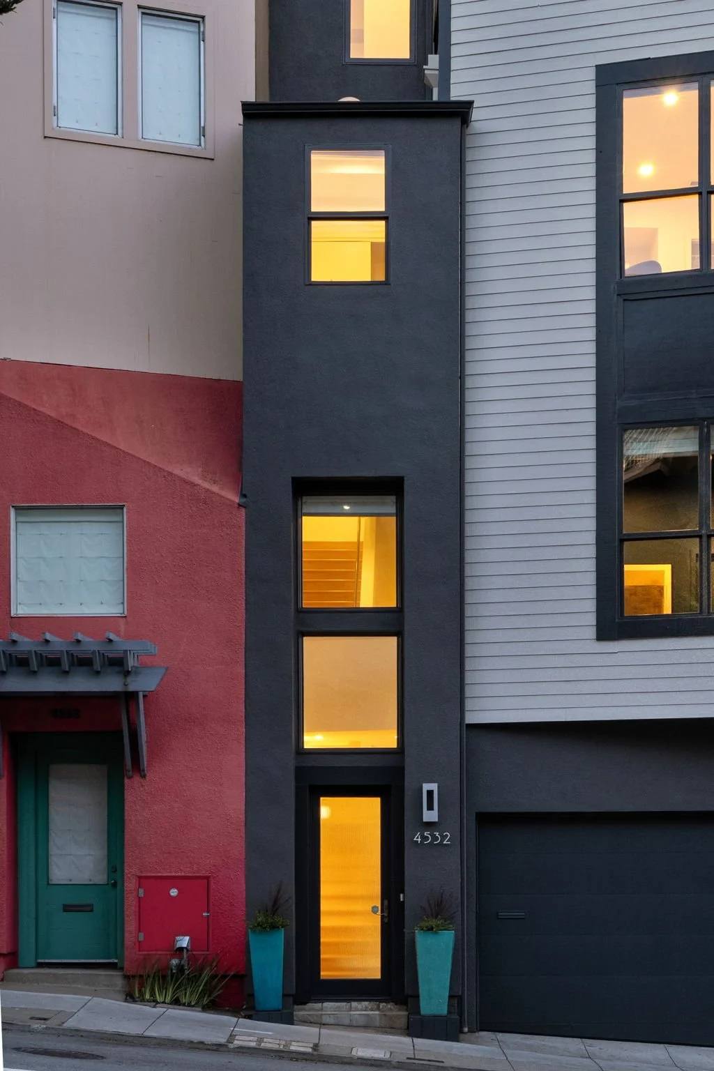 Modern multi-story residential building with yellow interior lights, a black front door, and two turquoise planters on either side of the entrance.