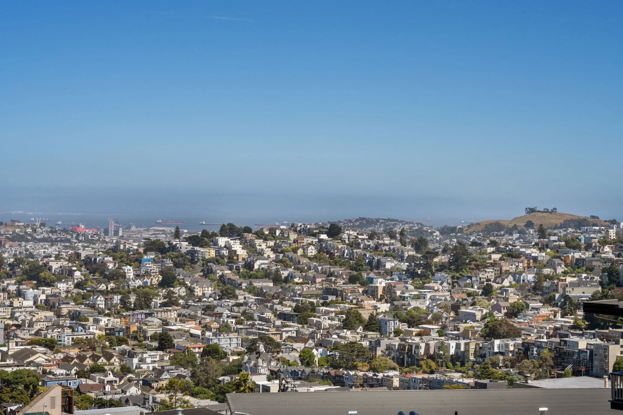 A panoramic view of a cityscape with numerous residential houses, a hill with sparse trees, and a distant bay with ships, under a clear blue sky.