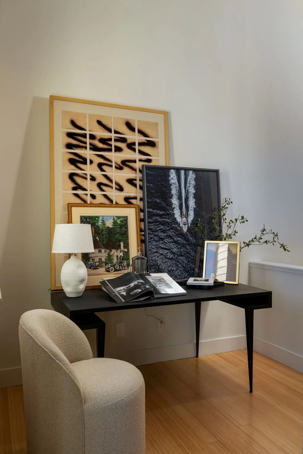 A black console table against a white wall holds framed photos, a white ceramic table lamp, a book, and a small potted plant with green leaves. Behind the table, there are large, framed art pieces, including a topographical map, a vintage house paint