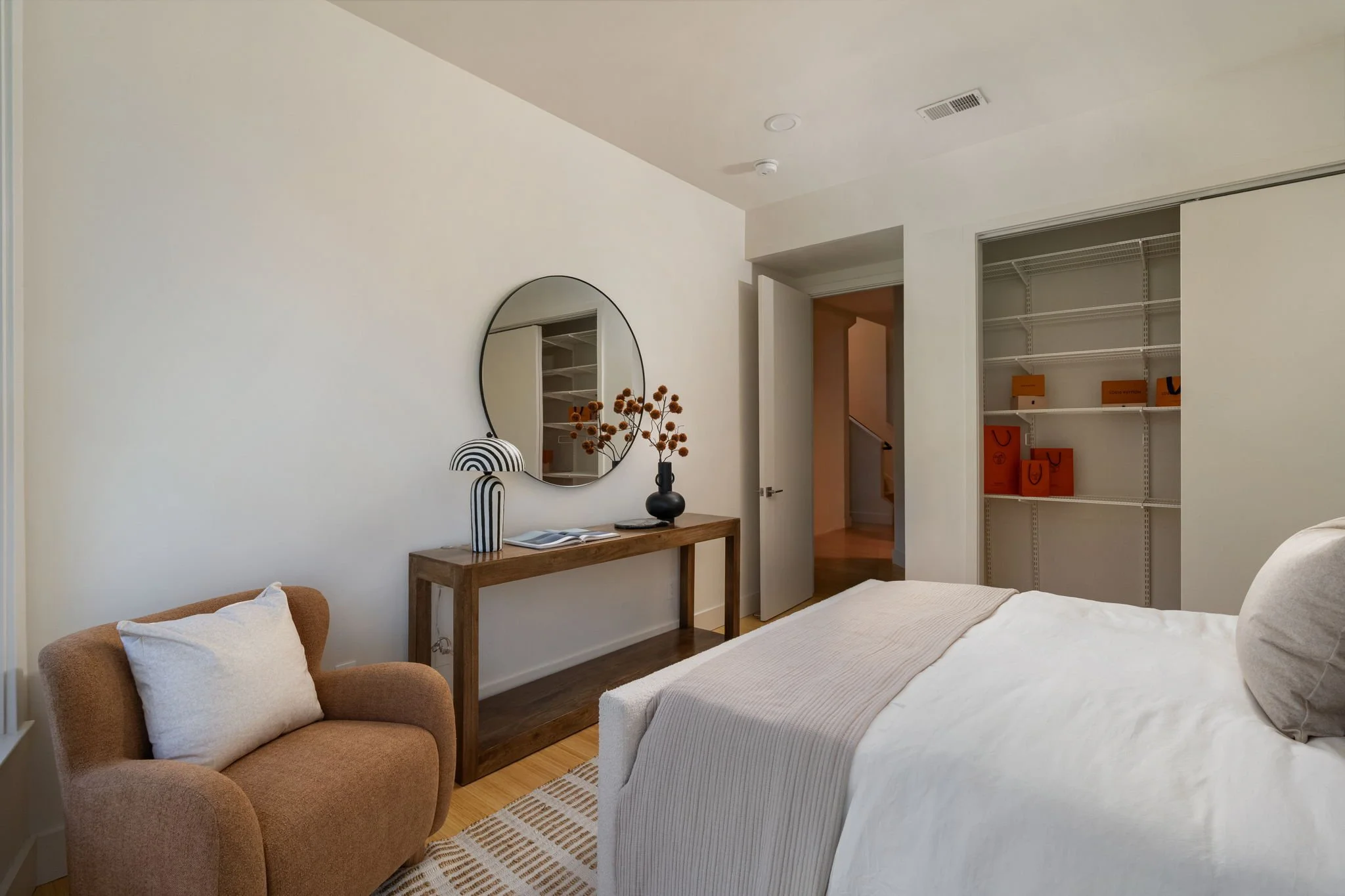 A bedroom with a bed, a side table, a mirror, and a corner shelf unit. The bed has white bedding and pillows. The side table has a black vase with dried flowers. A rounded armchair with white pillows is near the bed. The wall is painted white, and th