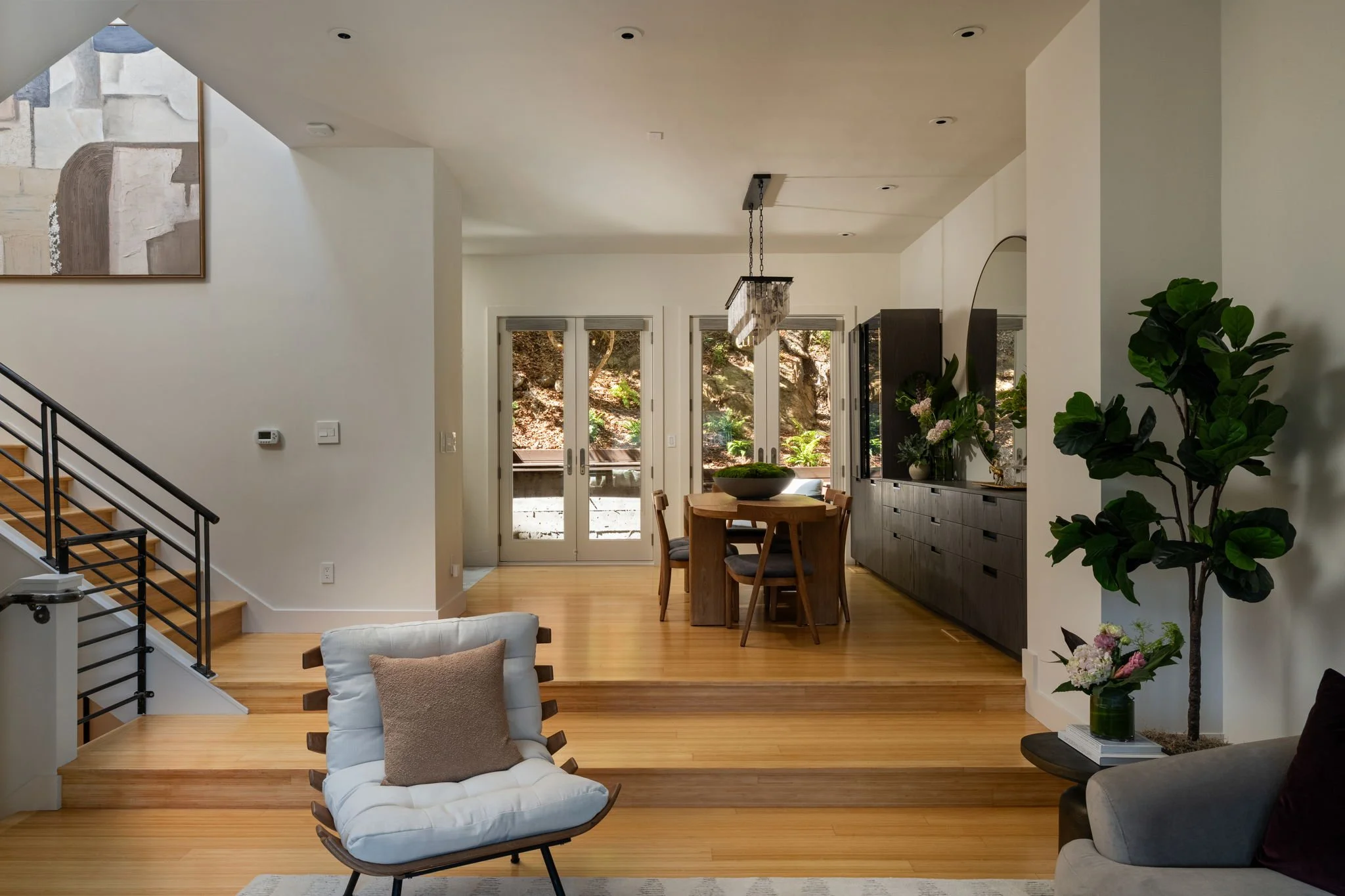 A spacious, modern living and dining area with sliding glass doors opening to an outdoor space with trees. The room has wooden flooring, a round dining table with chairs, a cozy lounge chair, and a large potted plant. There's artwork on the wall and a modern light fixture hanging above the dining area.