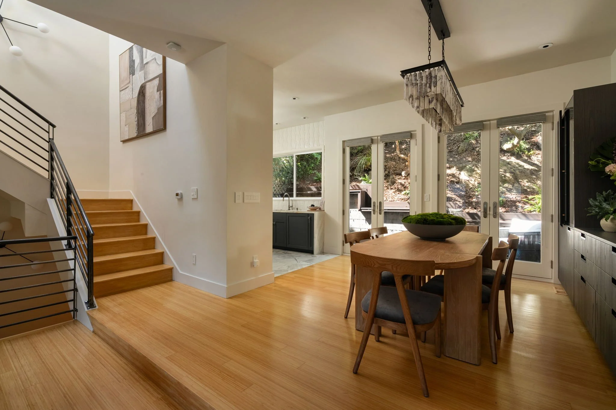 Open-concept living space with wooden flooring, a staircase with black metal railing on the left, a dining table with chairs, large windows with views of a hillside, and a kitchen area in the background.