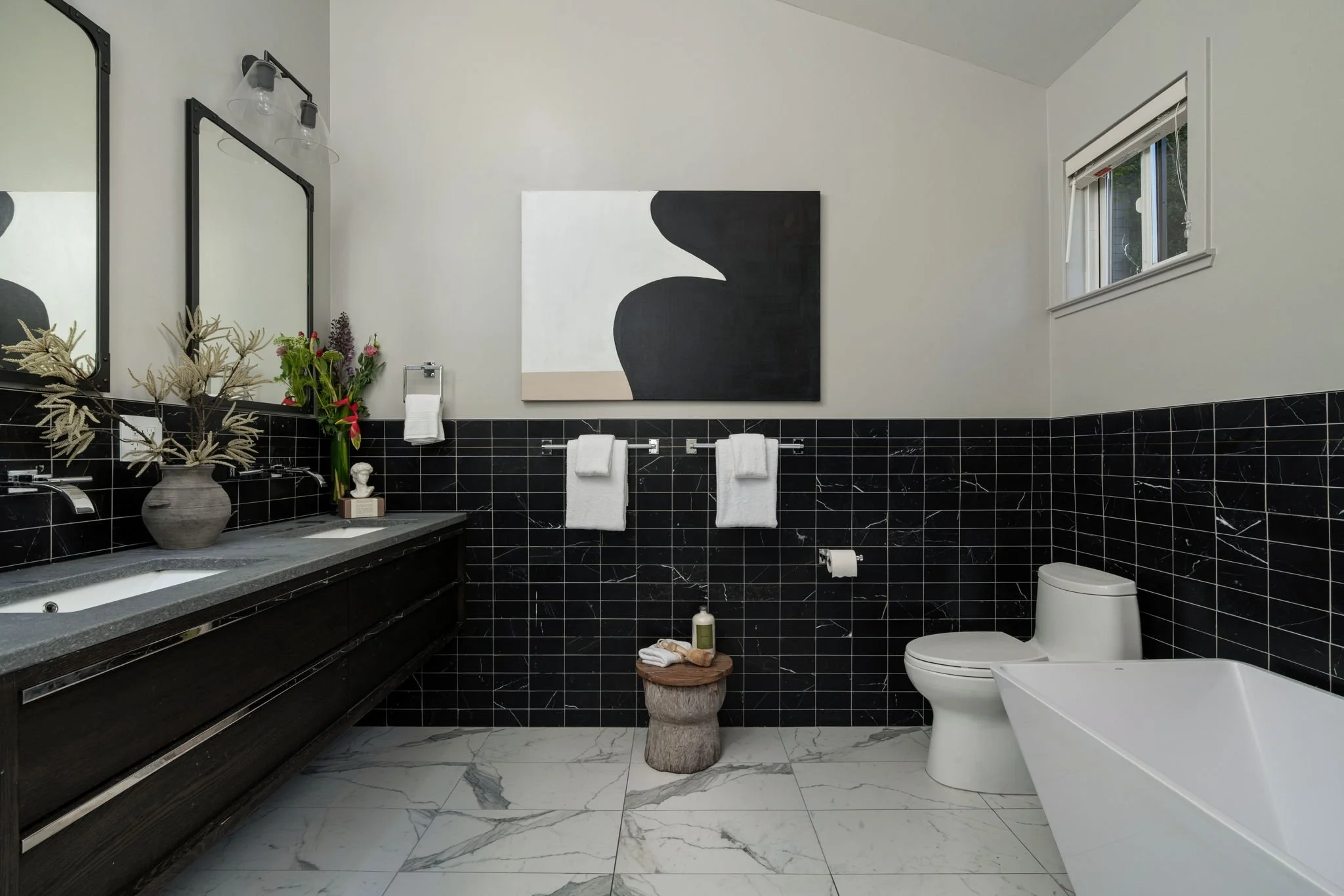 A modern bathroom with white walls and black marble lower walls. There are two fridges, a mirror, a vase with flowers, and a towel holder. The bathroom has a small window with blinds, a white toilet, and some toiletries on a small table.