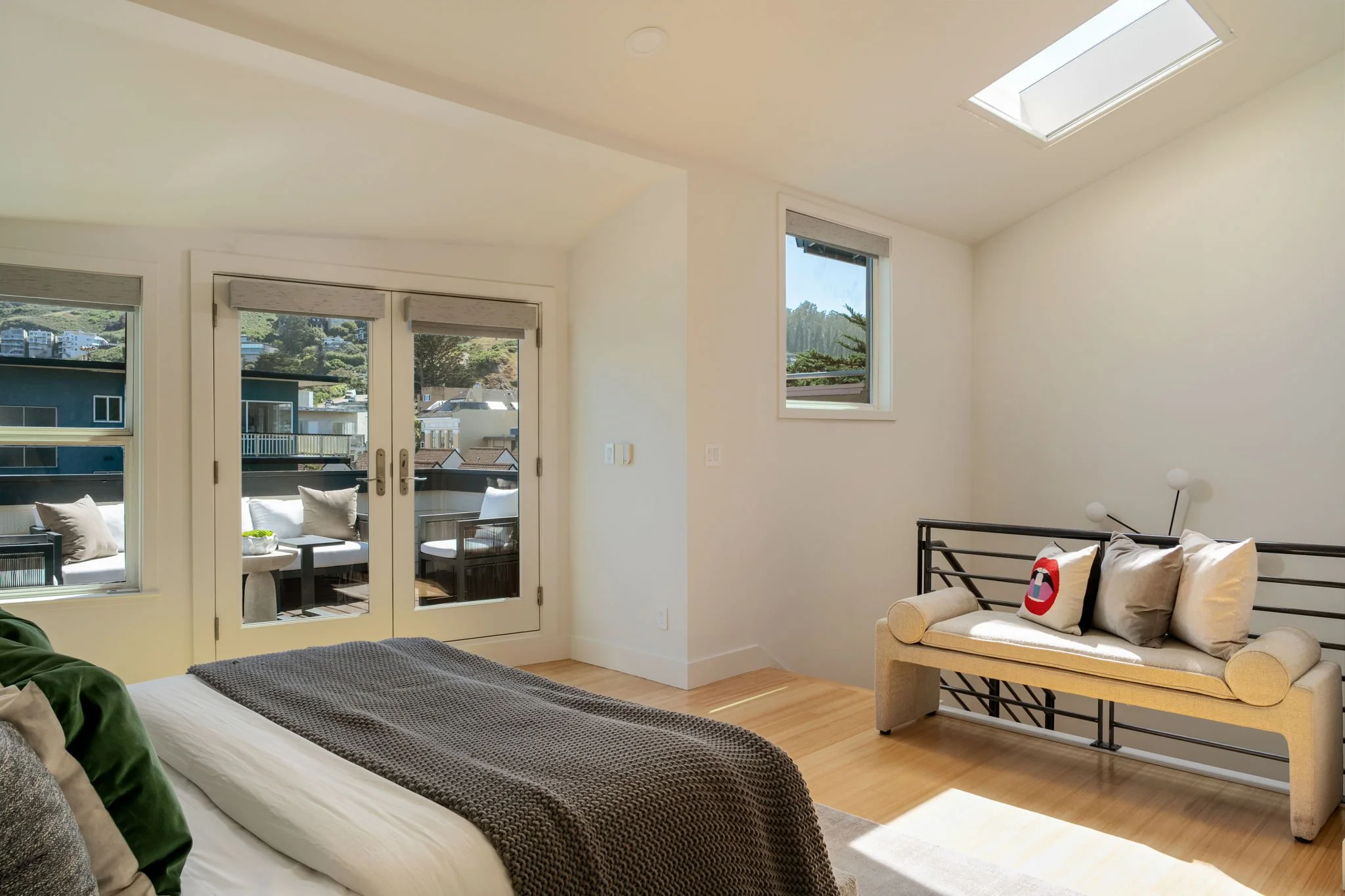 A bright modern living room with large glass doors and windows, white walls, and wooden flooring. There is a patio with outdoor seating visible through the doors, and a skylight in the ceiling.