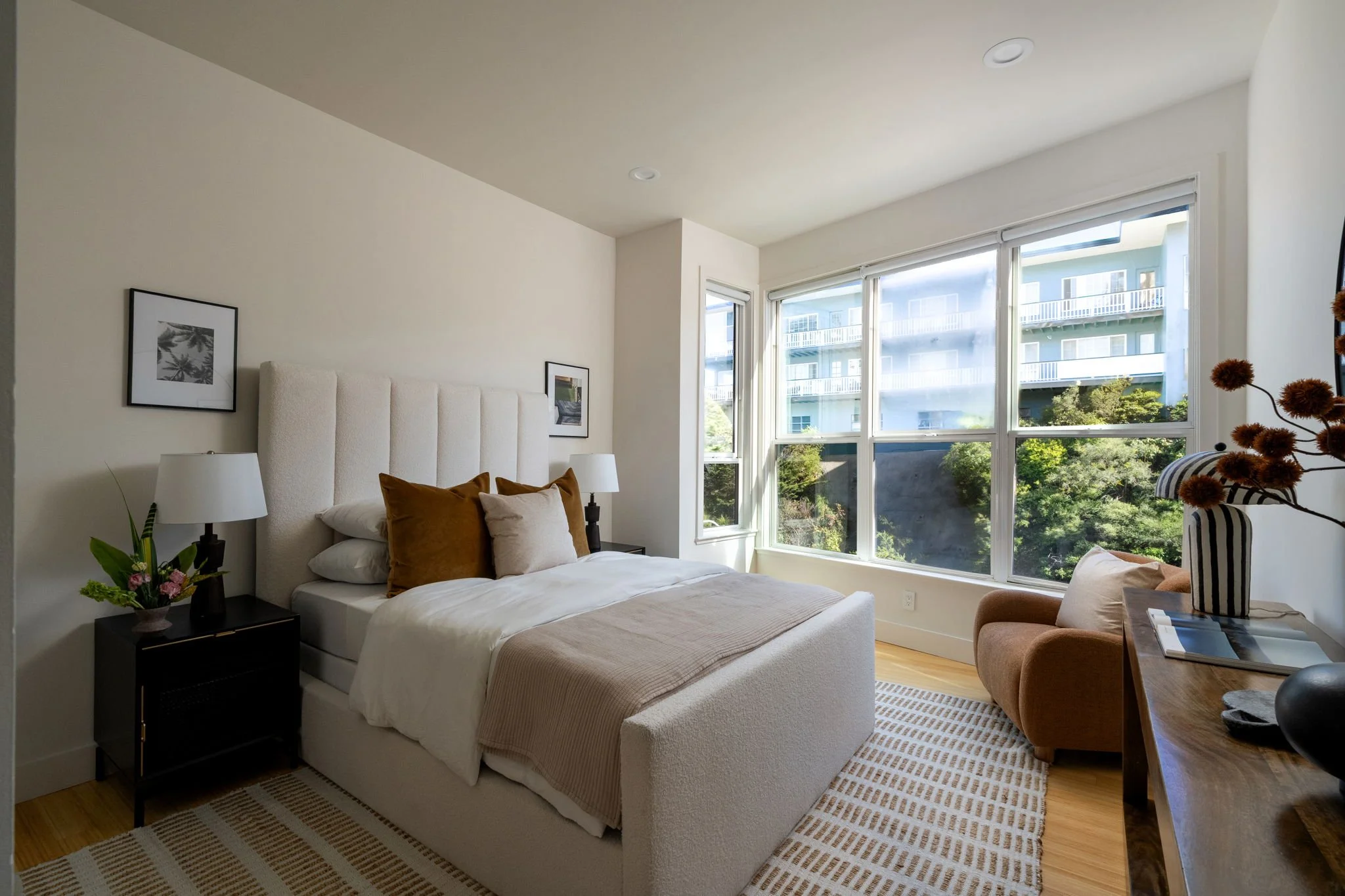 A bedroom with a large window showing nearby residential buildings and trees, white bed with brown and cream pillows, two black nightstands with lamps and flowers, and an armchair with striped pillows and a side table.