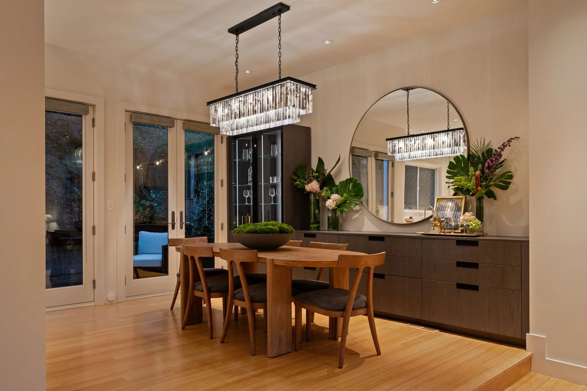 A modern dining room with a wooden table surrounded by chairs, a round mirror on the wall, a black display cabinet with glass shelves, and hanging rectangular chandelier lights. There are large sliding glass doors with beige blinds and a view of an o