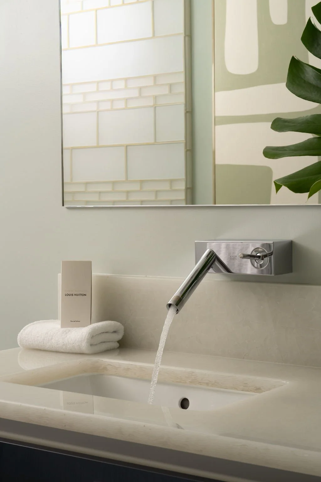 Bathroom sink with a modern metallic faucet, a white rolled towel, and a Louis Vuitton card. A large mirror and a green plant are visible in the background.