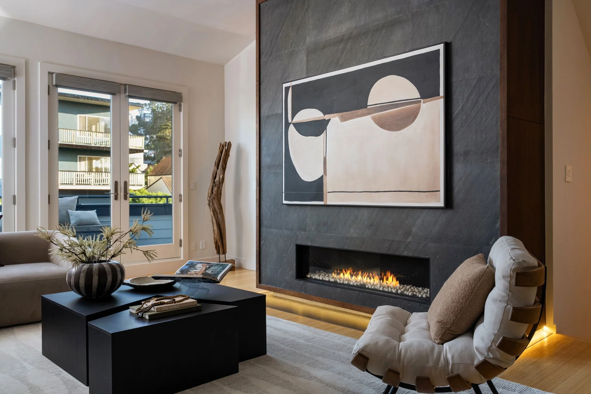 Living room with a modern fireplace, abstract wall art, beige sofa, black coffee table, glass door, and outdoor view of balconies and trees.