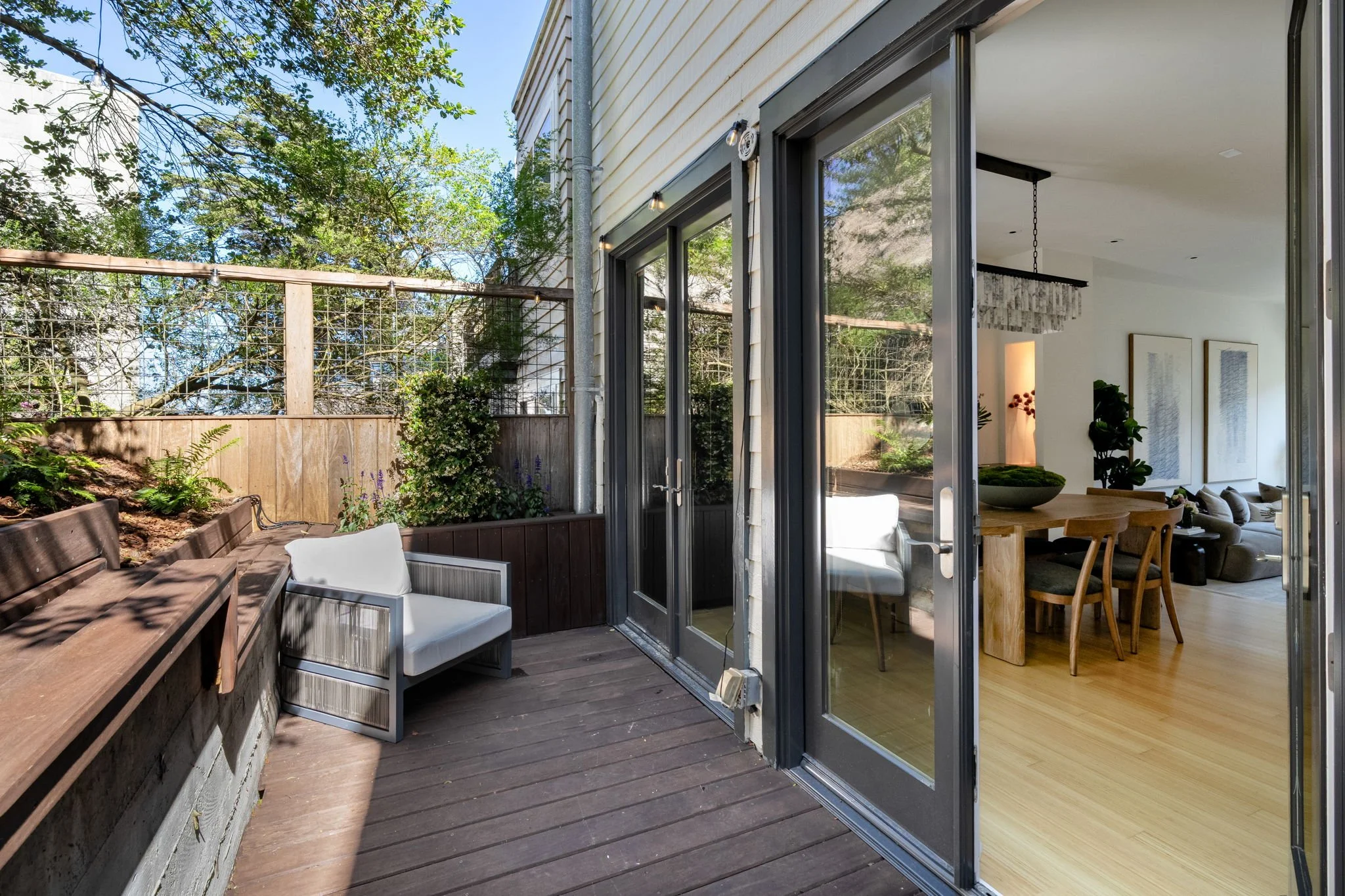 A backyard patio with wooden flooring, outdoor seating including a bench and an armchair, and a garden area with plants, enclosed by a wooden fence with string lights, adjacent to a house with large glass doors leading inside.