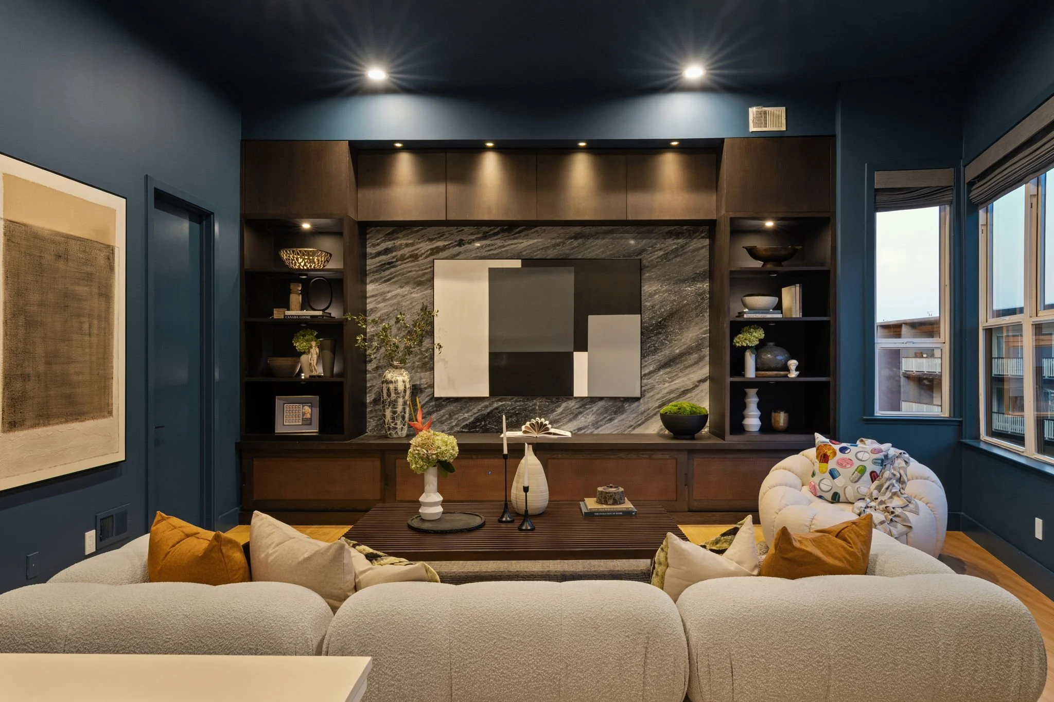 Living room with blue walls, dark wood built-in shelves, abstract art, and a marble fireplace. There are beige sofas with colorful pillows, a dark wood coffee table with decorative items, and windows with gray blinds.