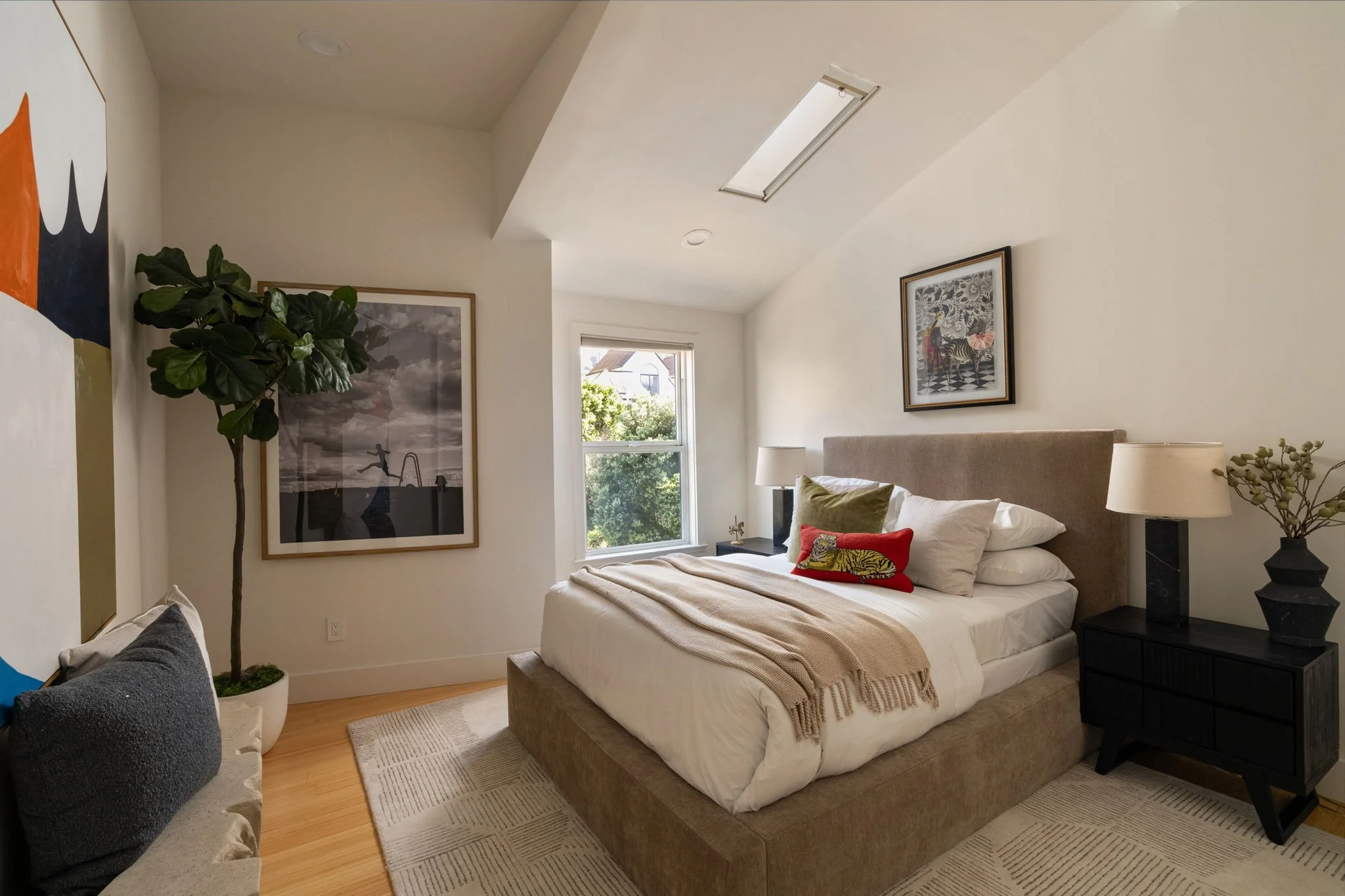 A cozy bedroom with a large bed with white bedding and decorative pillows, dark nightstands with lamps, framed artwork on cream-colored walls, a window with a view of greenery outside, a house, and ceiling skylight.