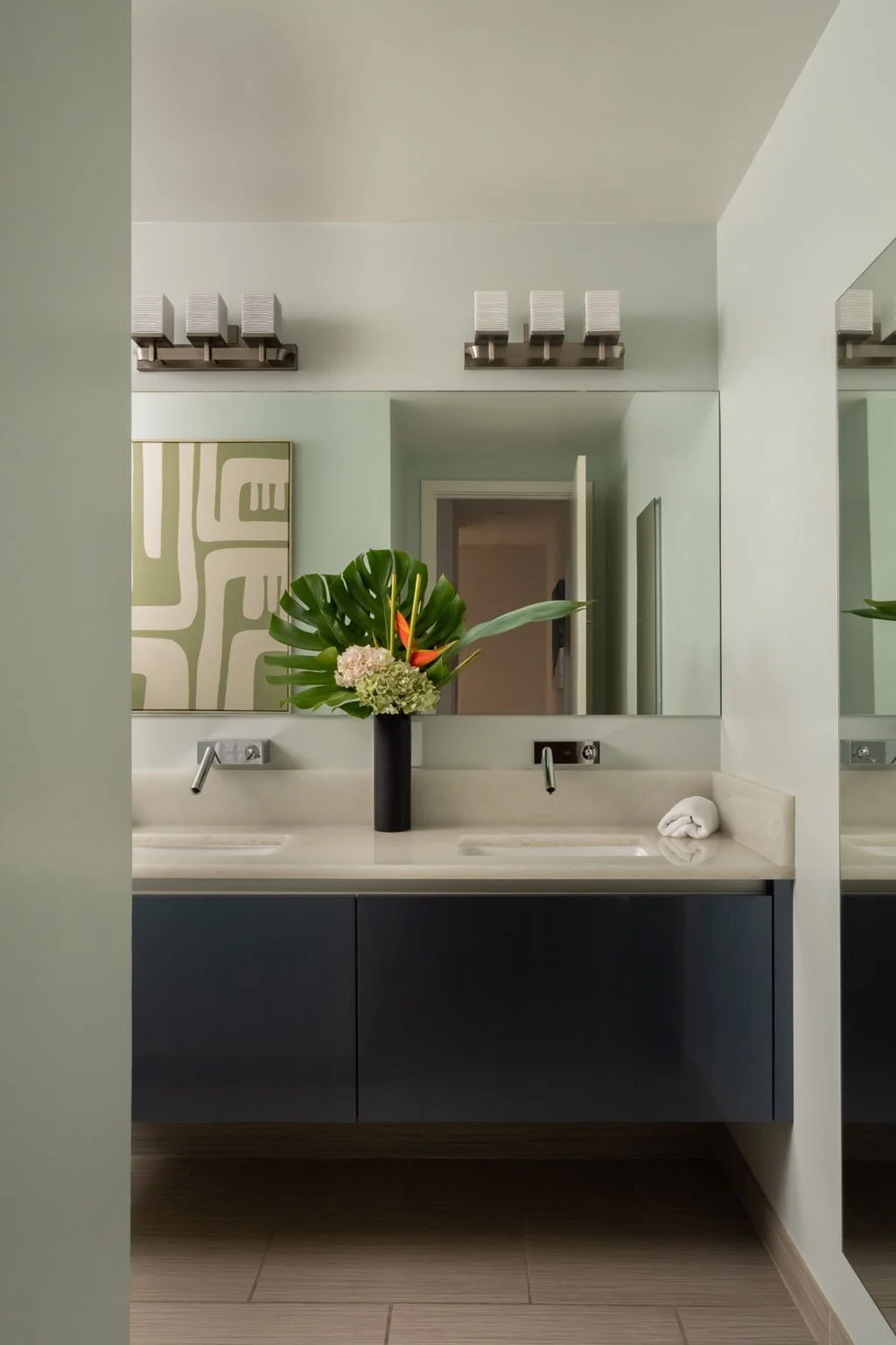 Bathroom with a large mirror, a black vase with tropical flowers and greenery on the sink, a rolled white towel, and modern light fixtures above.