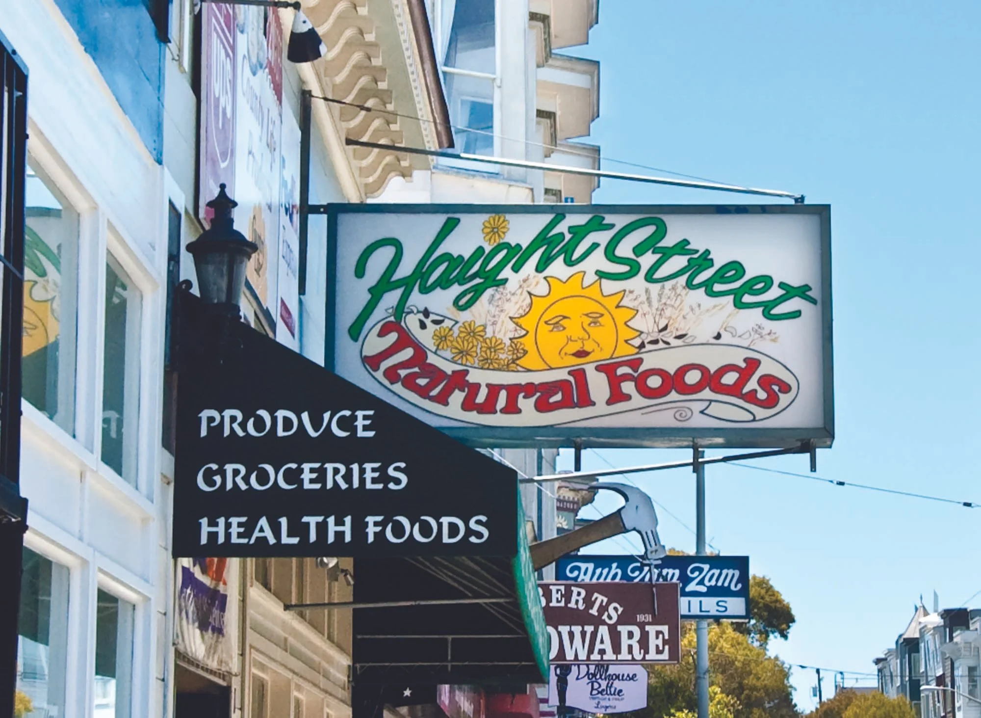 Street view showing multiple shop signs, including Haag Street Natural Foods and a black sign with white text for groceries and health foods. Buildings line the street with blue sky overhead.